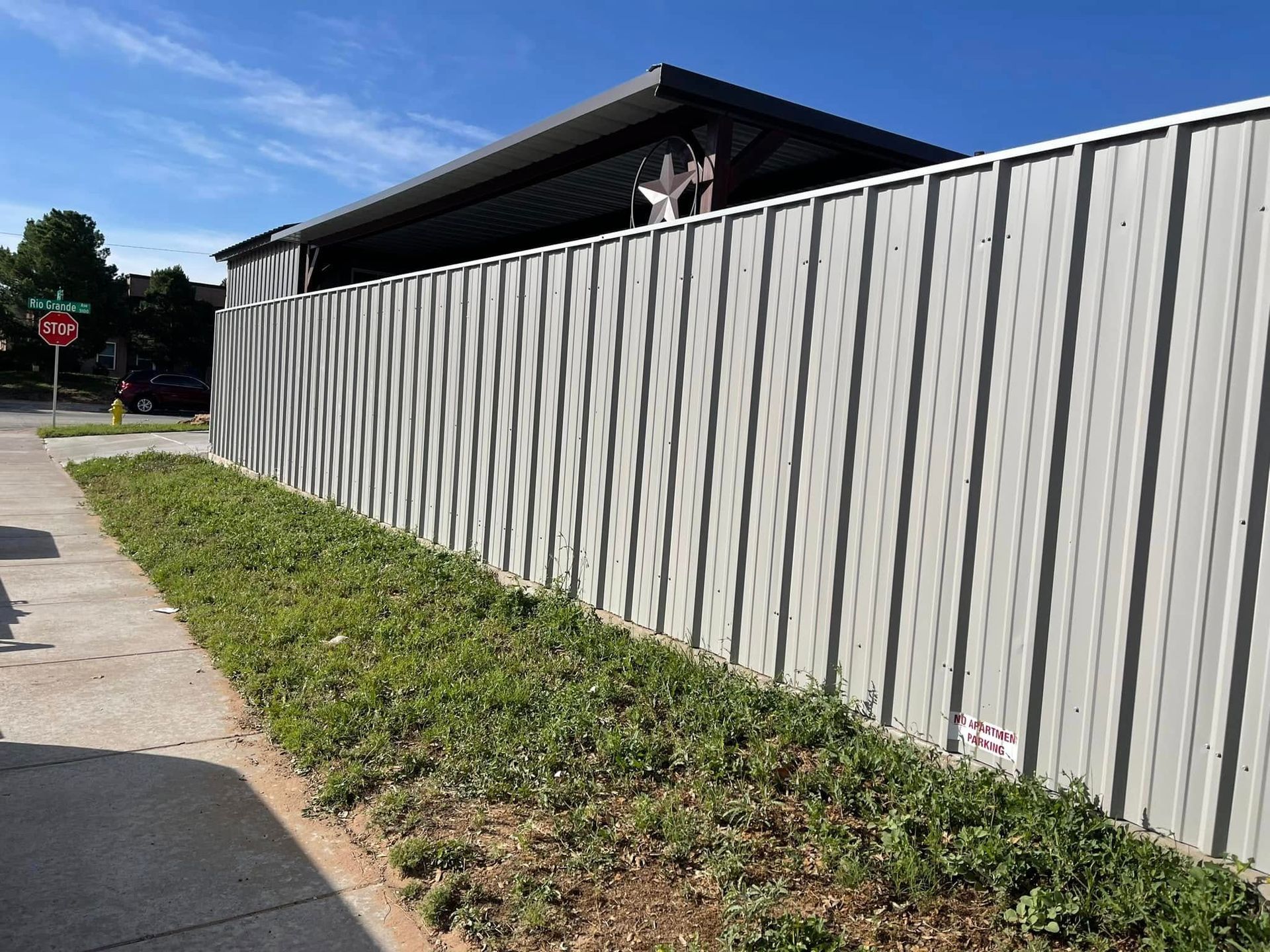 Person peeking over a tall gray fence beside a sidewalk and grassy verge
