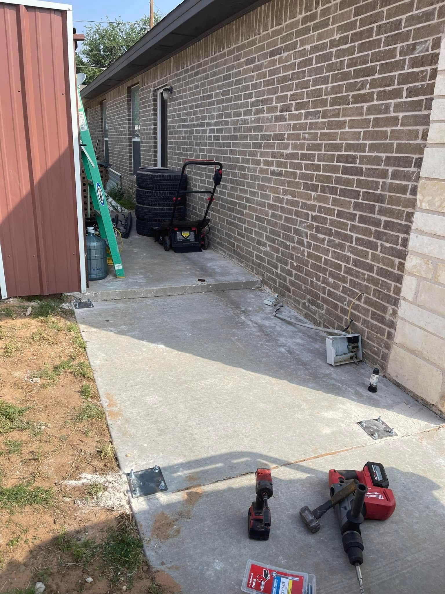 Narrow concrete side yard beside a brick house, with tools and a wheelbarrow near the wall.