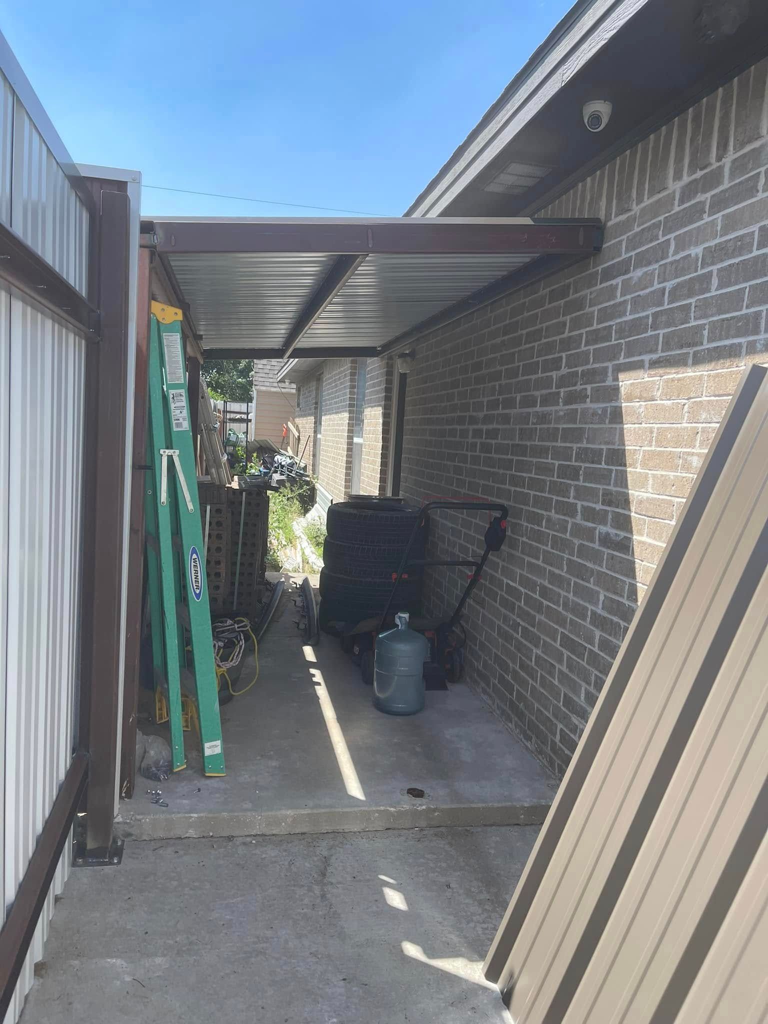 Narrow side yard with brick house wall, metal awning, stacked panels, and gardening tools.