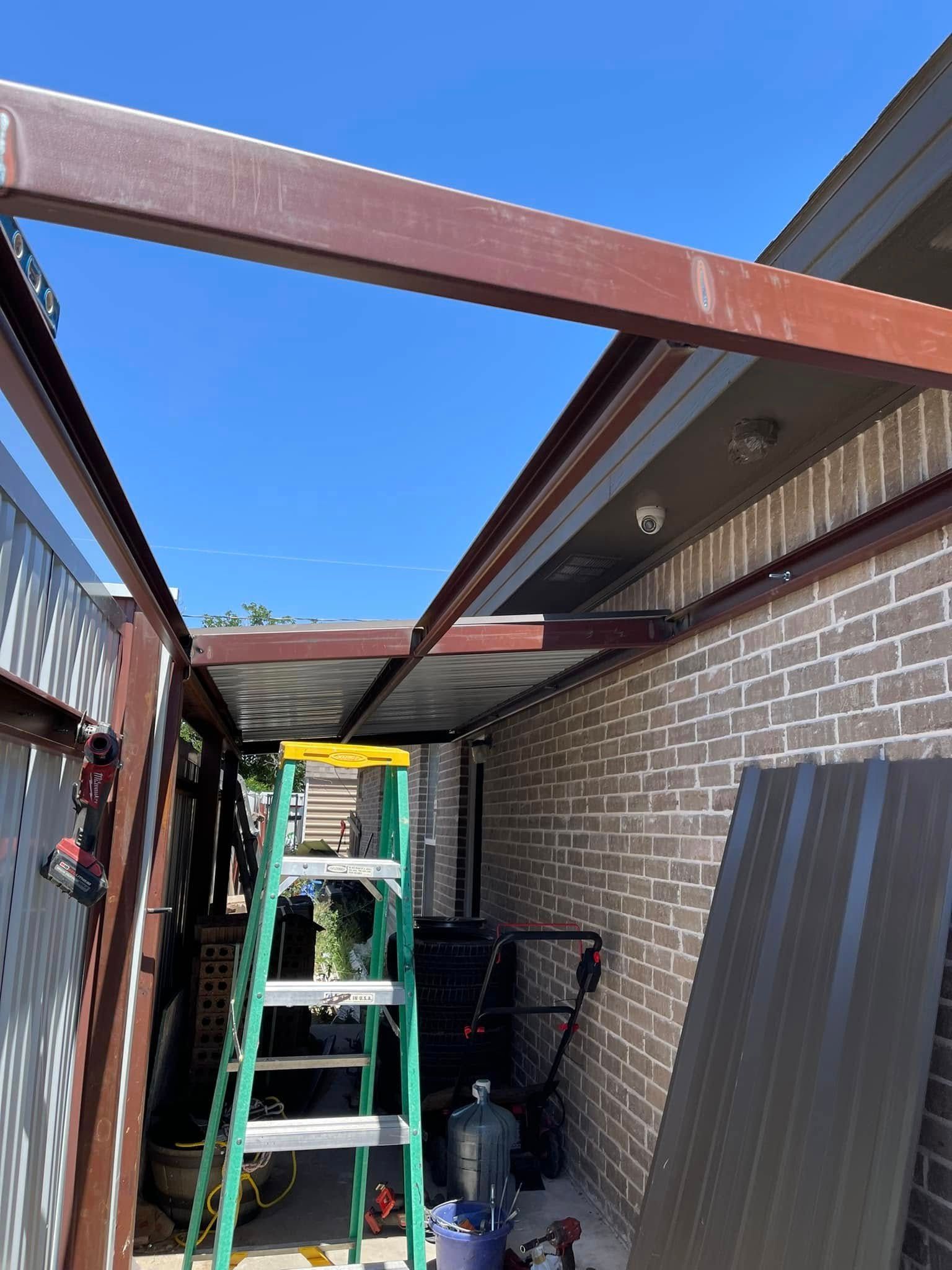 Covered side yard with metal beams, ladder, and construction materials beside a brick house.