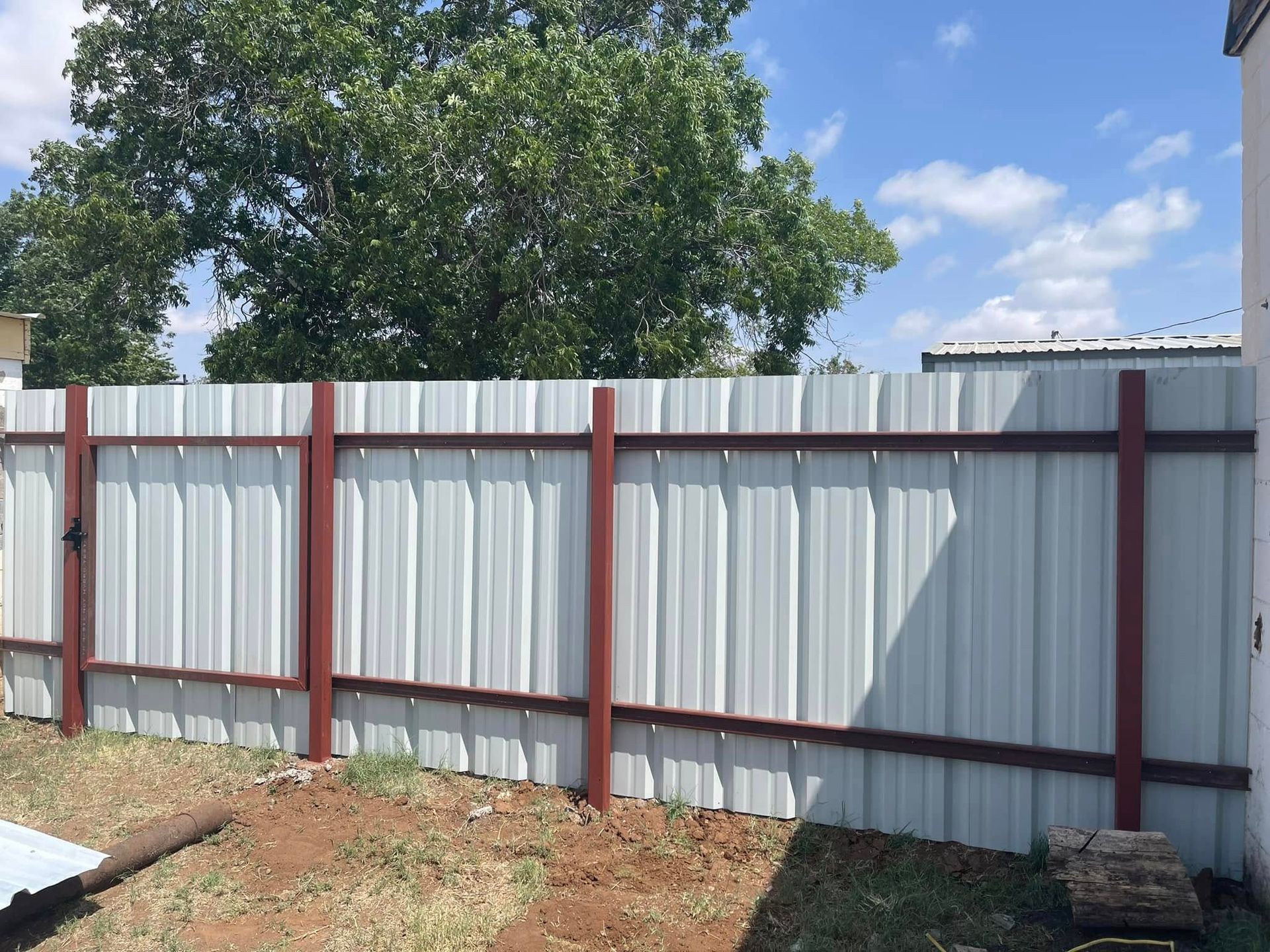 Corrugated metal fence with red posts in a yard, trees and blue sky behind it.