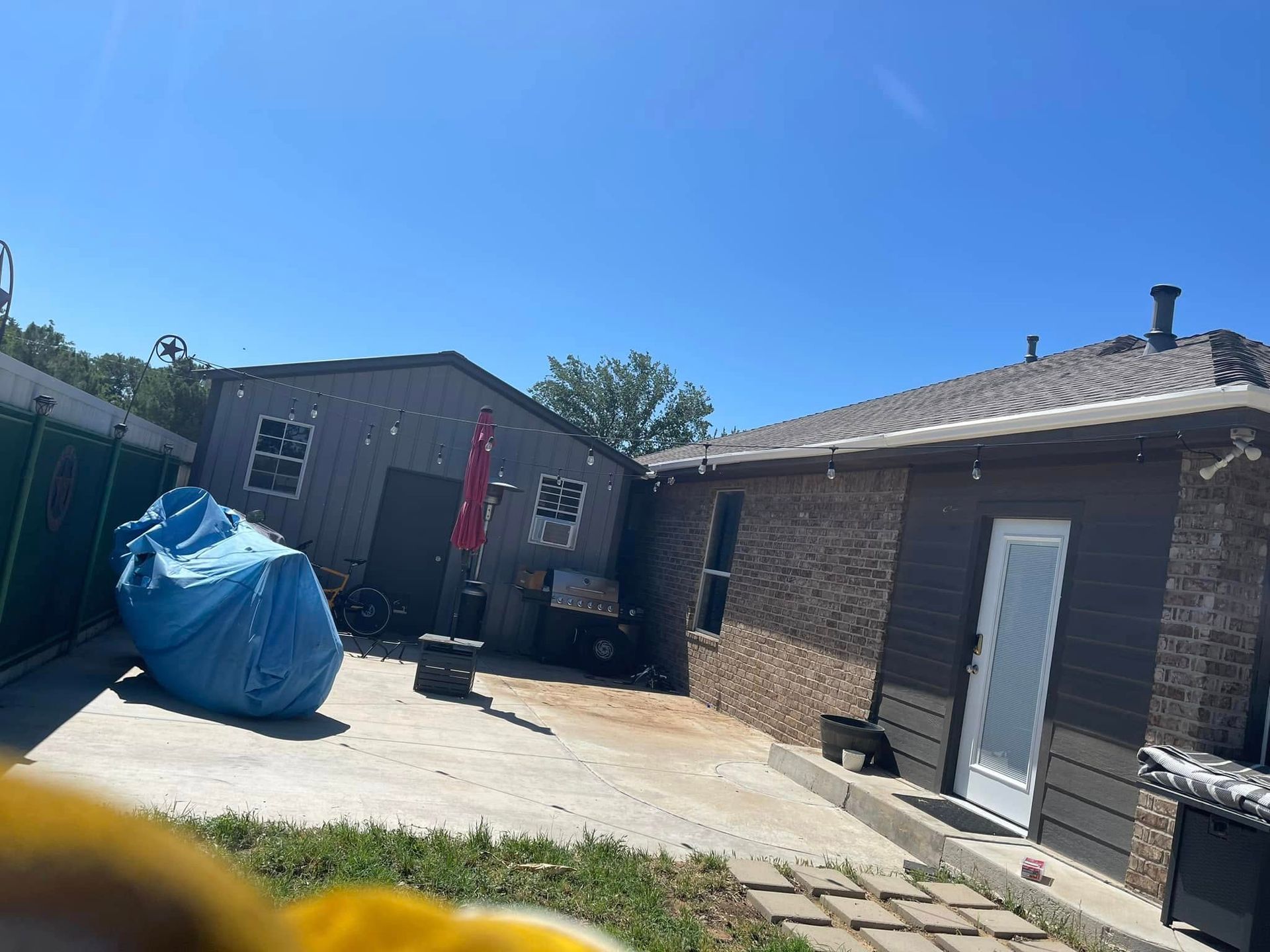 Backyard patio with brick house, gray shed, blue tarp-covered item, and black grill under a clear sky