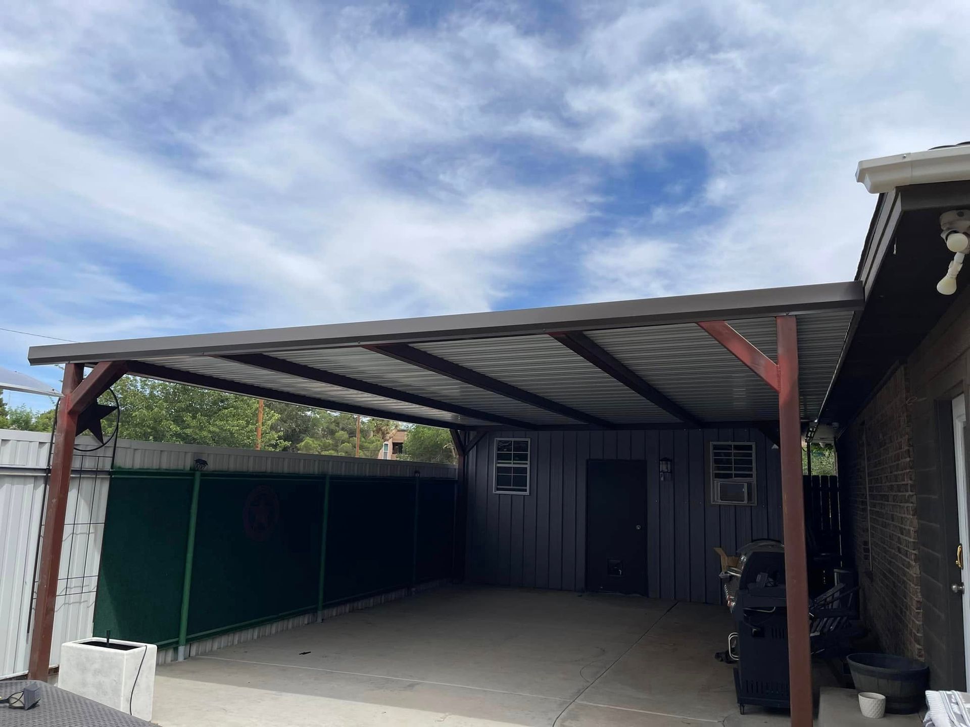 Covered patio/carport beside a house with a green fence and cloudy sky