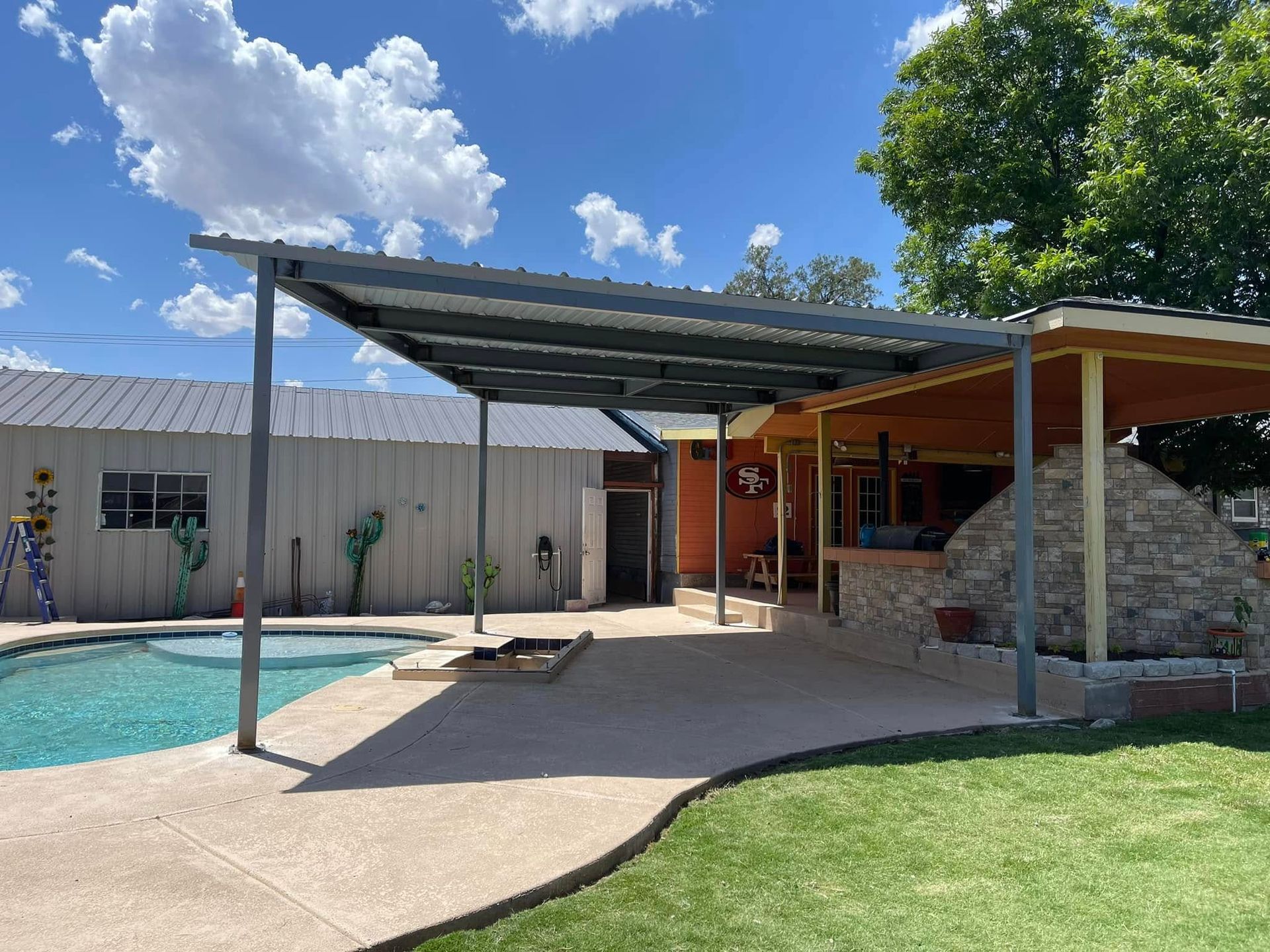 Backyard pool beside a covered patio and stone fire feature under a bright blue sky