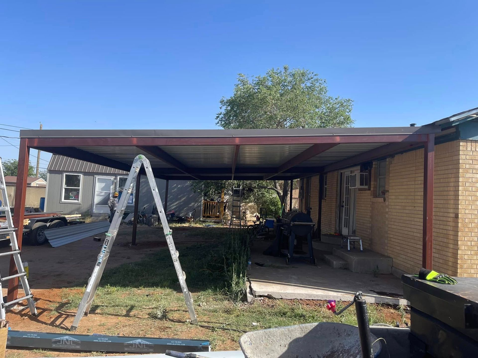 Carport under construction beside a brick house, with a ladder and scattered tools in the yard.