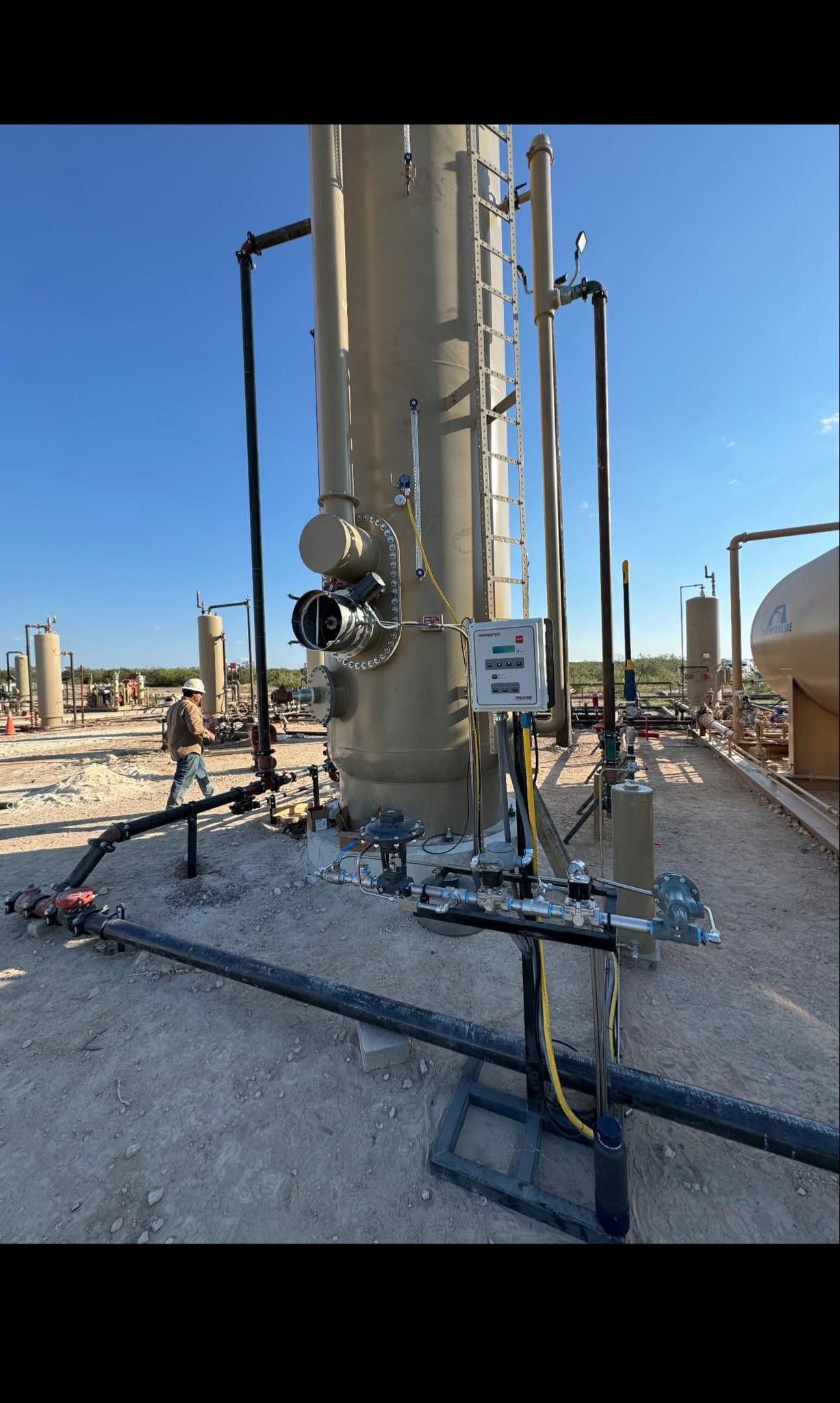 Industrial refinery equipment with tall metal tank, pipes, valves, and hoses in a sunny outdoor yard