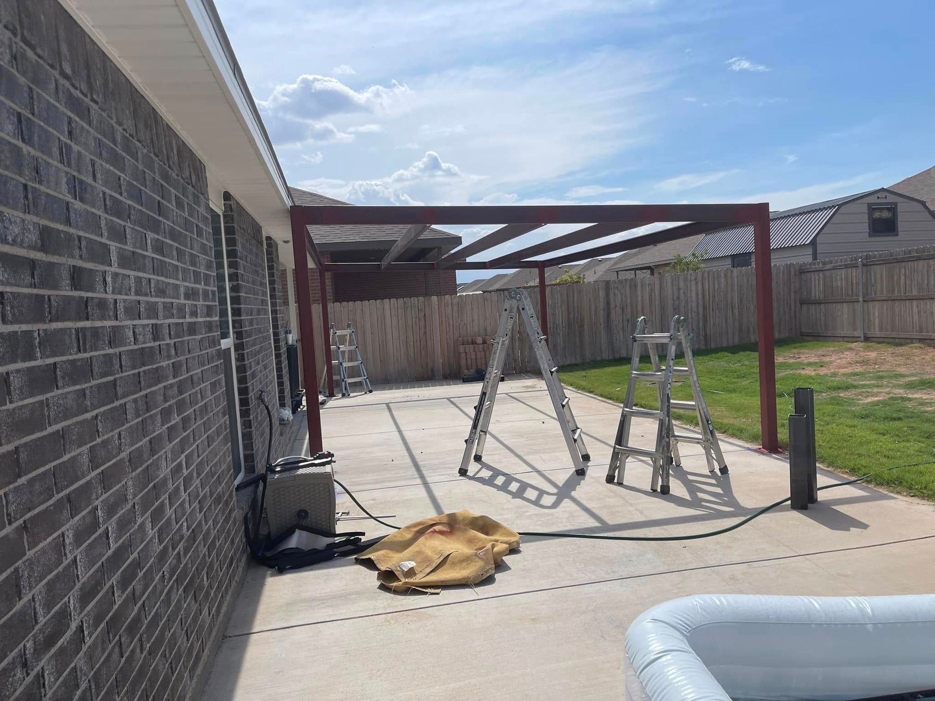 Backyard patio with a metal pergola frame, ladders, and construction tools beside a brick house