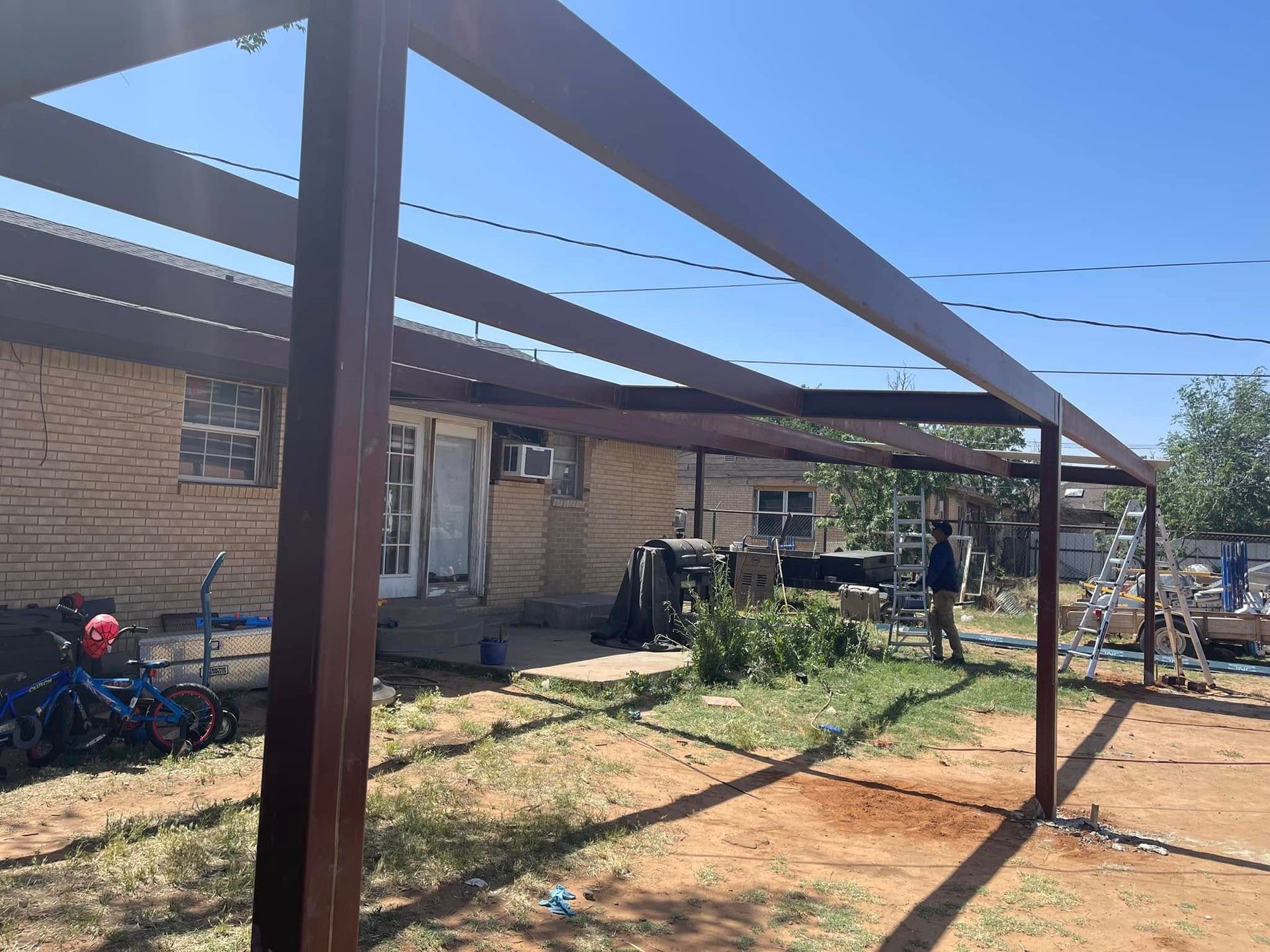 Metal frame structure over a dirt yard beside a brick building, with bicycles and a person in the background.