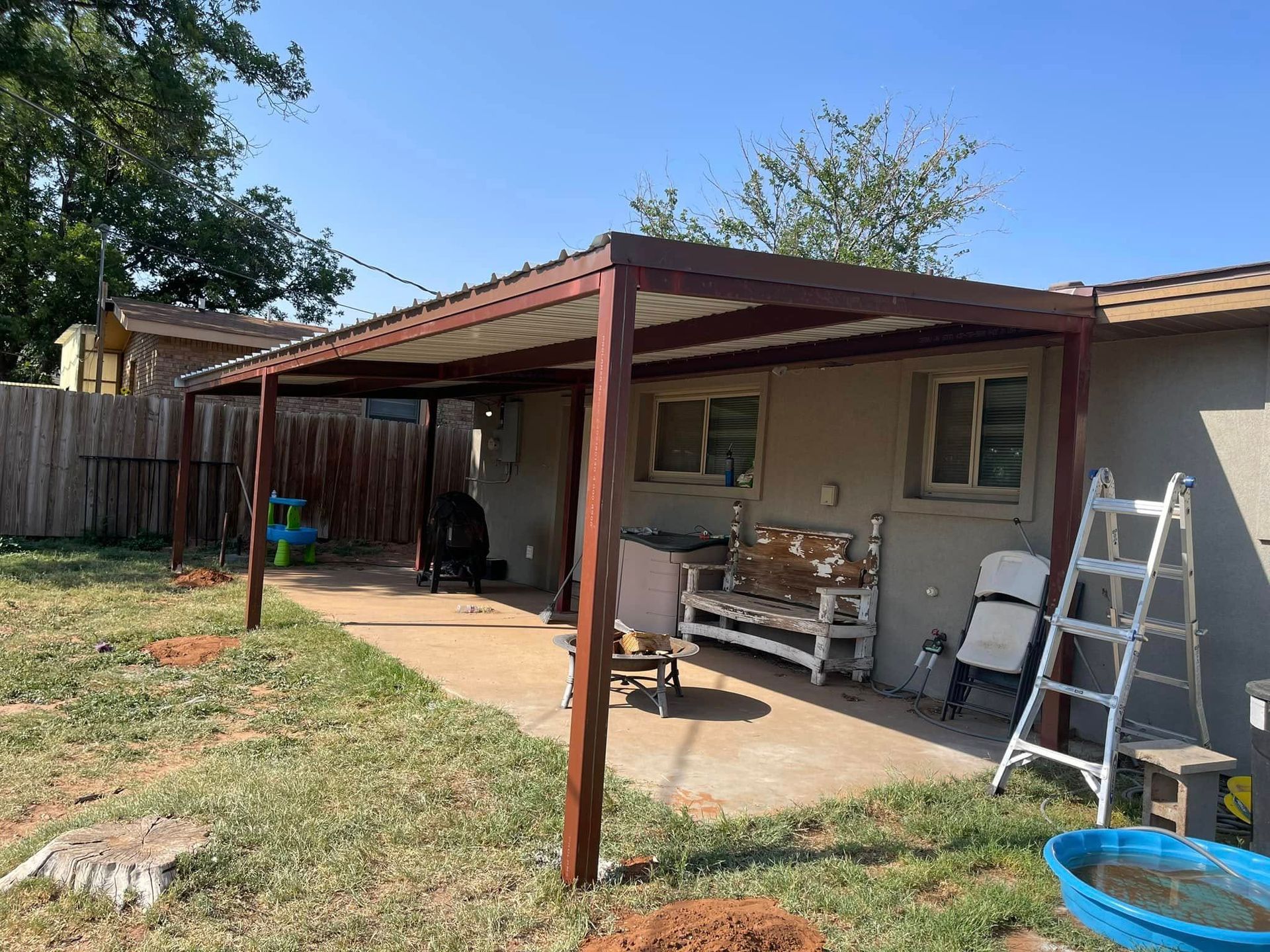 Backyard patio with red pergola, ladder, bench, and workbench beside a house.
