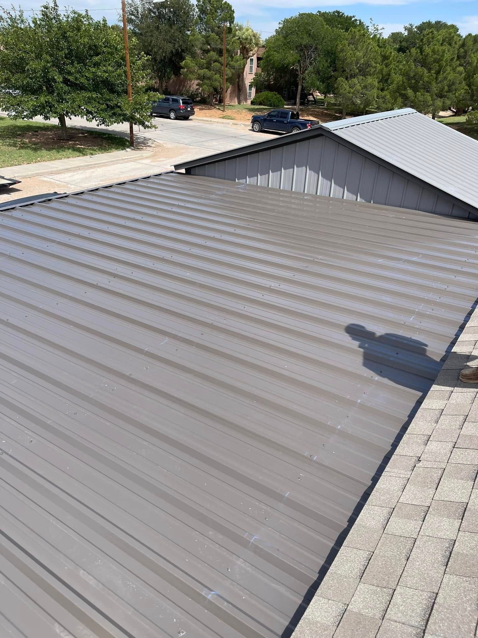Gray corrugated metal roof beside a small gabled roof, with trees and parked cars in the background.