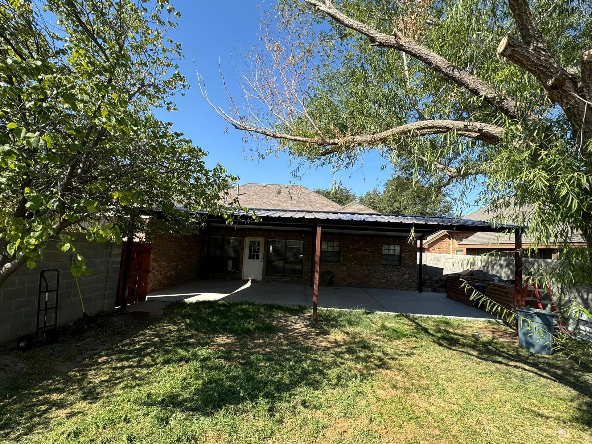 Backyard with green lawn, shaded patio, and trees under a clear blue sky