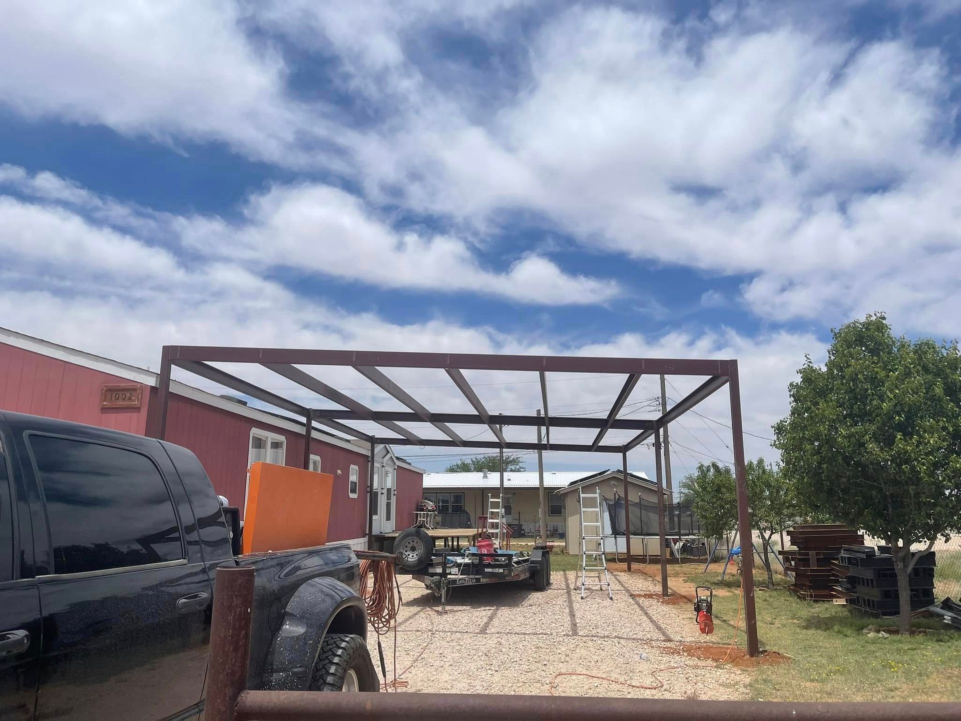 Metal carport frame in a gravel driveway with a truck and van under a cloudy blue sky