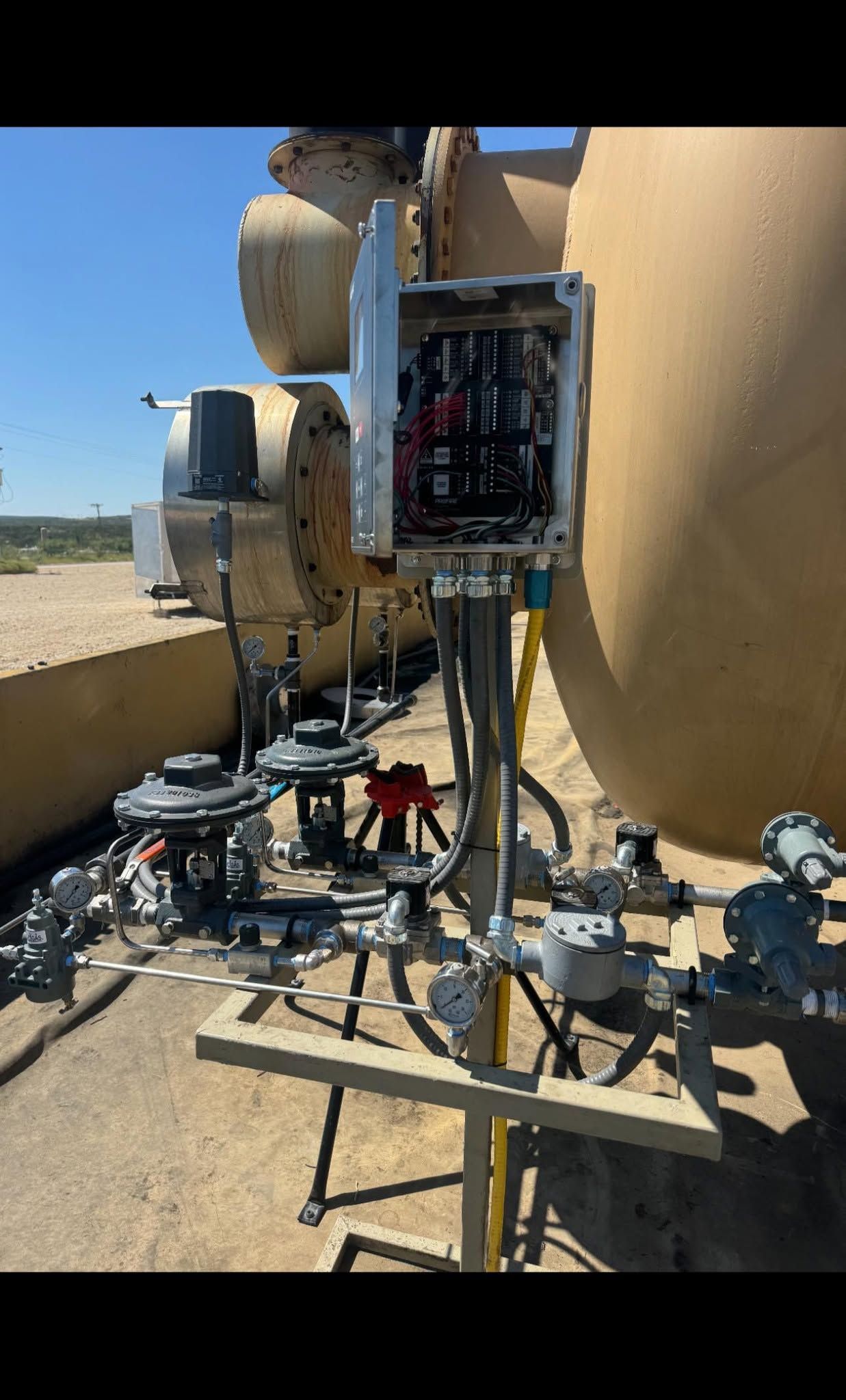 Electrical control panel beside beige industrial tanks and metal piping outdoors in bright sun