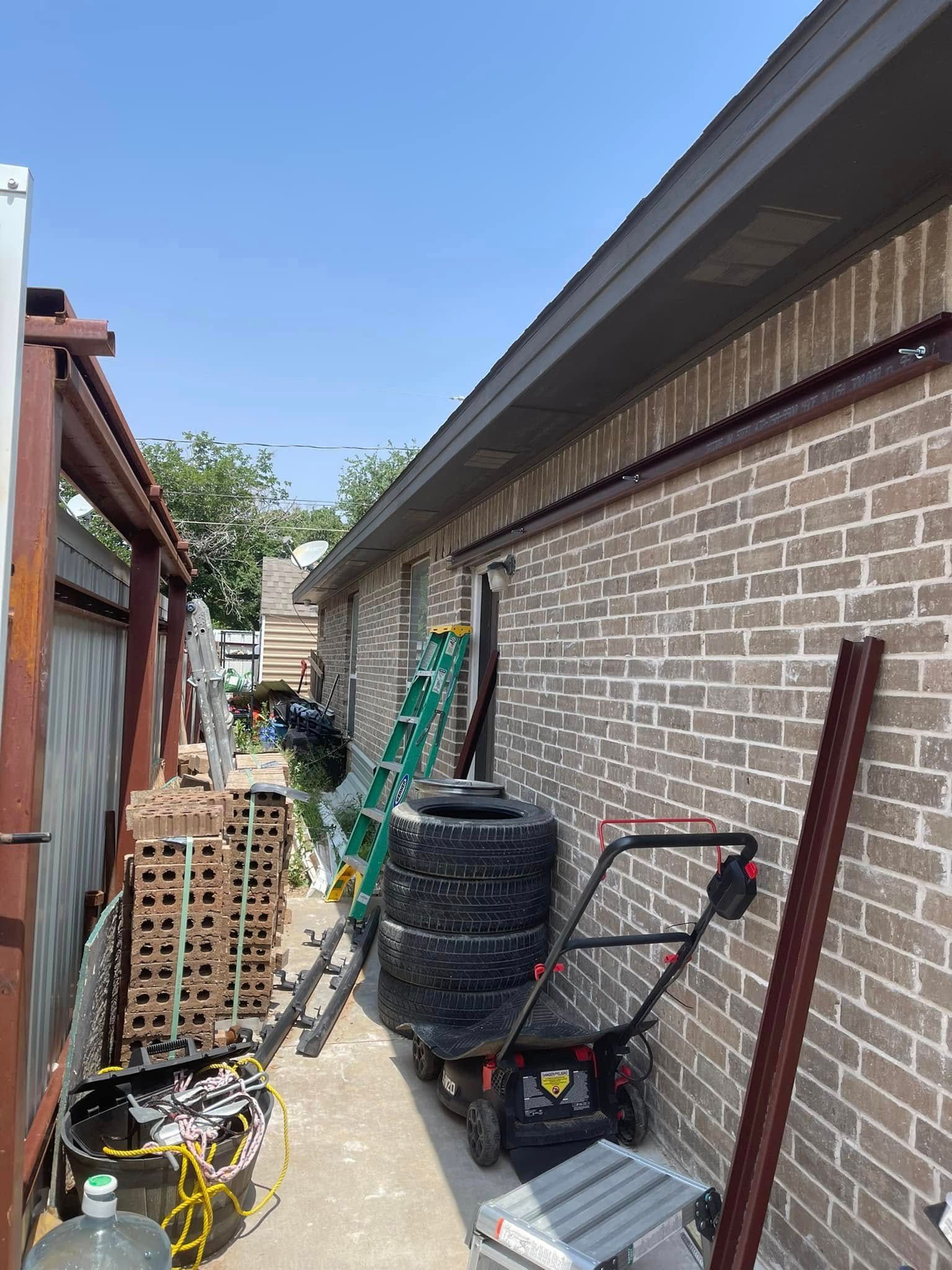 Narrow outdoor alley beside a brick building, crowded with stacked pallets, tools, and a black water barrel.