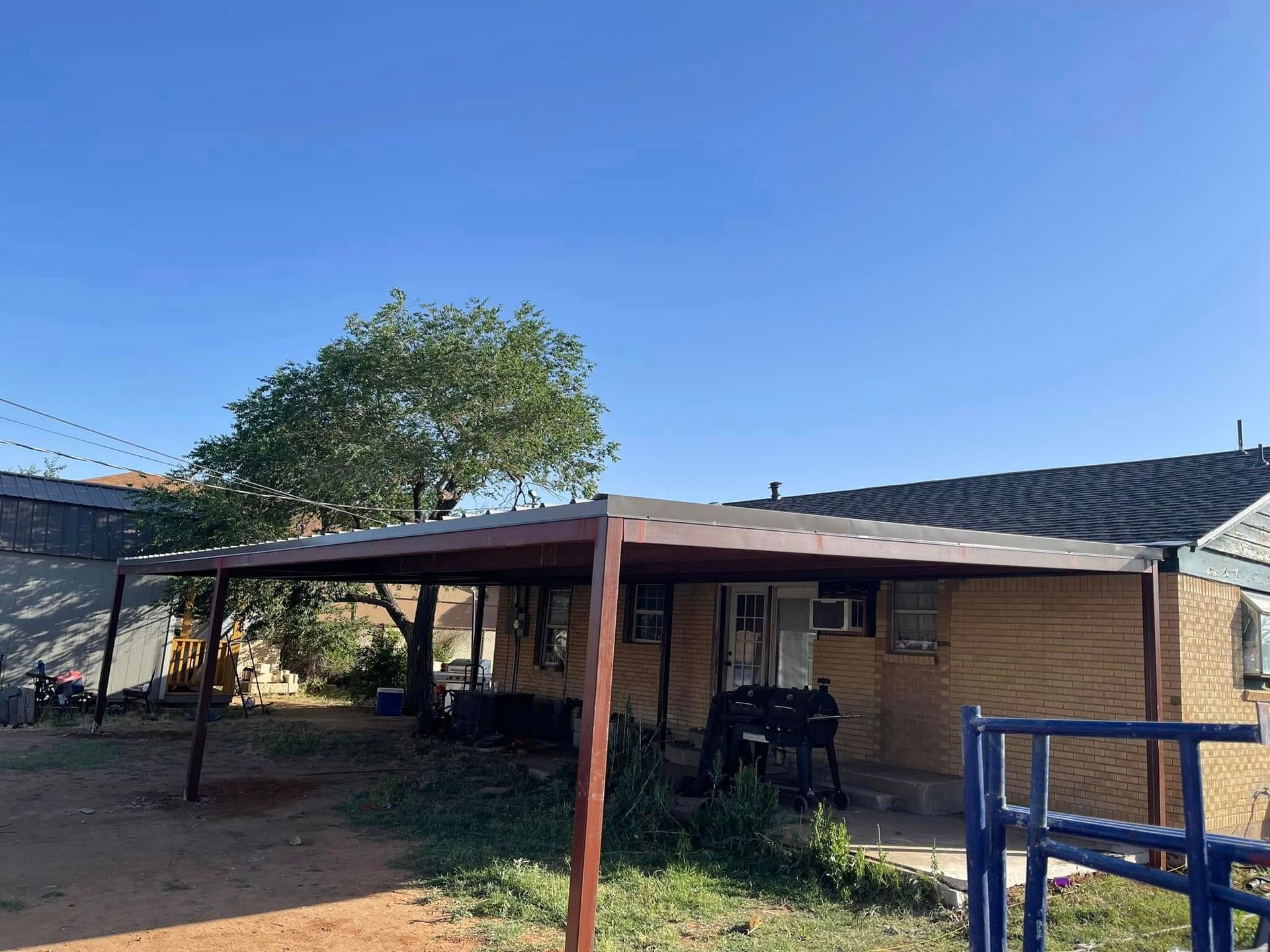 Rural house with a shaded carport, dirt yard, and a blue gate under a clear sky