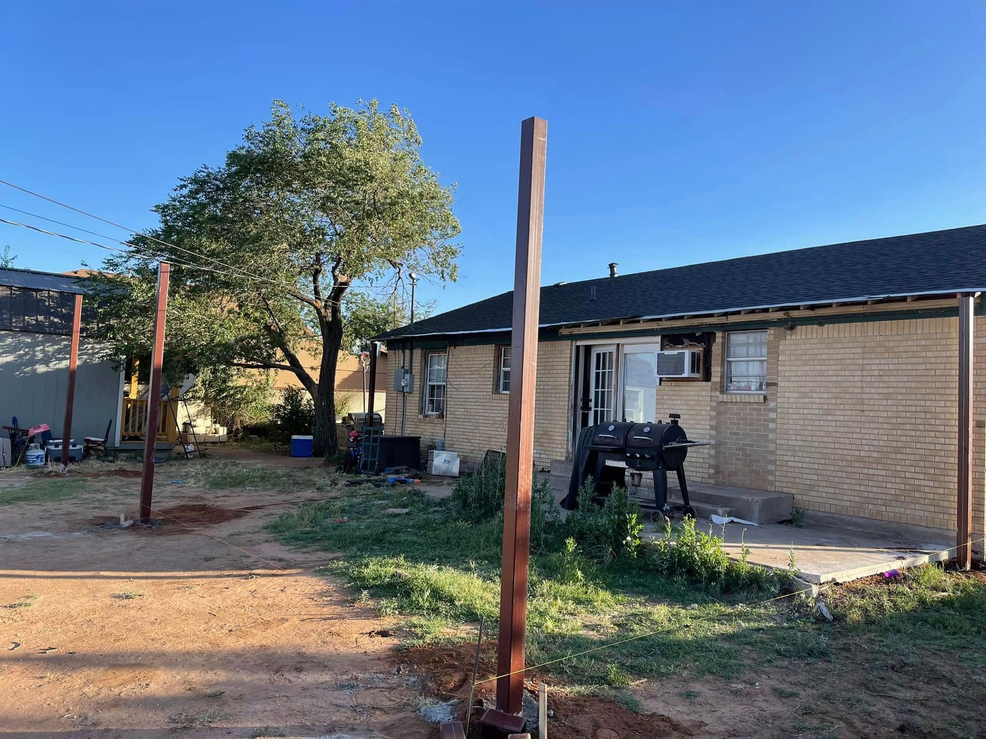 Backyard with dirt ground, wooden posts, a tree, and a brick house with a patio chair under a clear blue sky