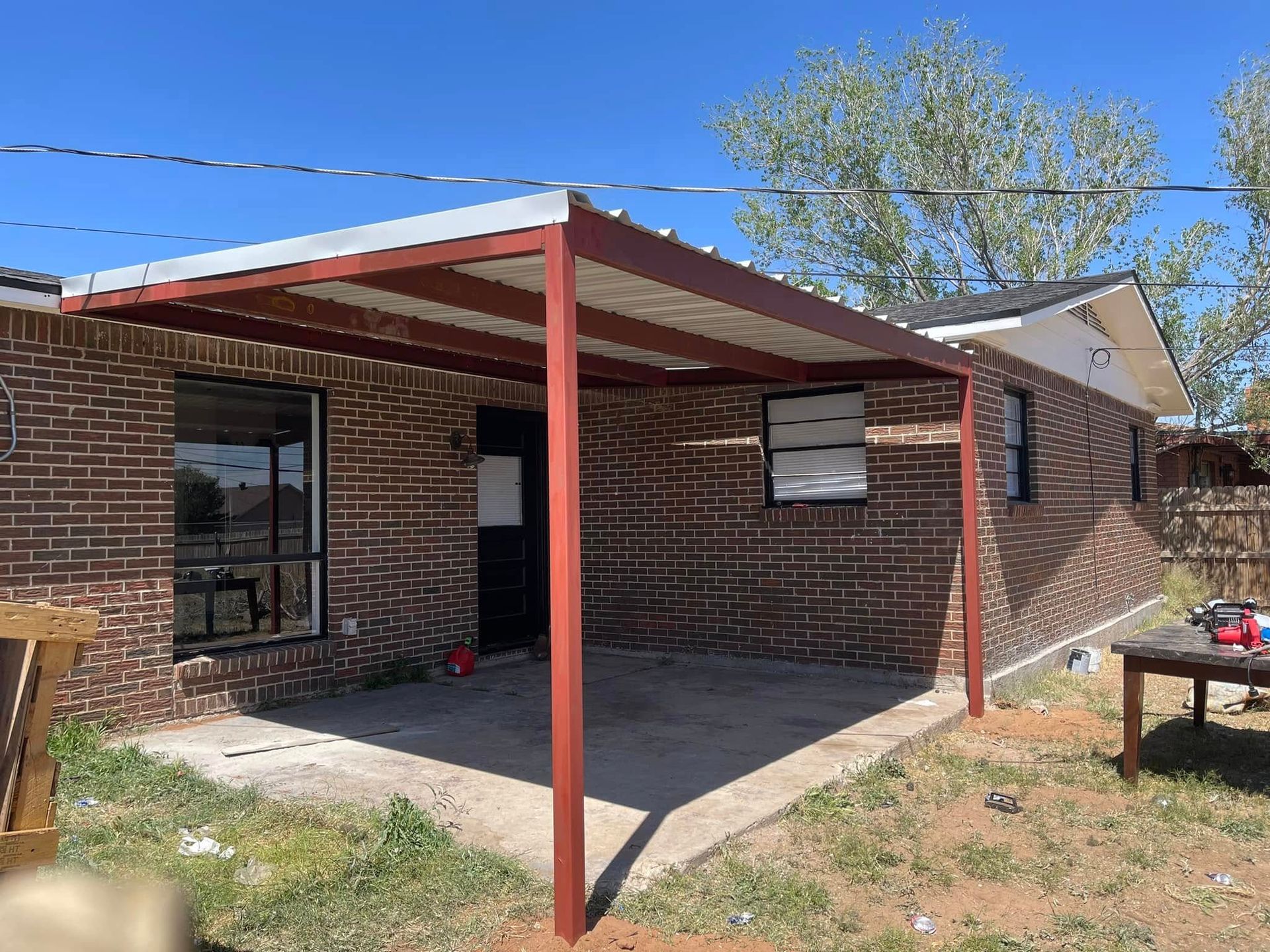 Covered brick patio with red posts and a concrete slab beside a house under a blue sky
