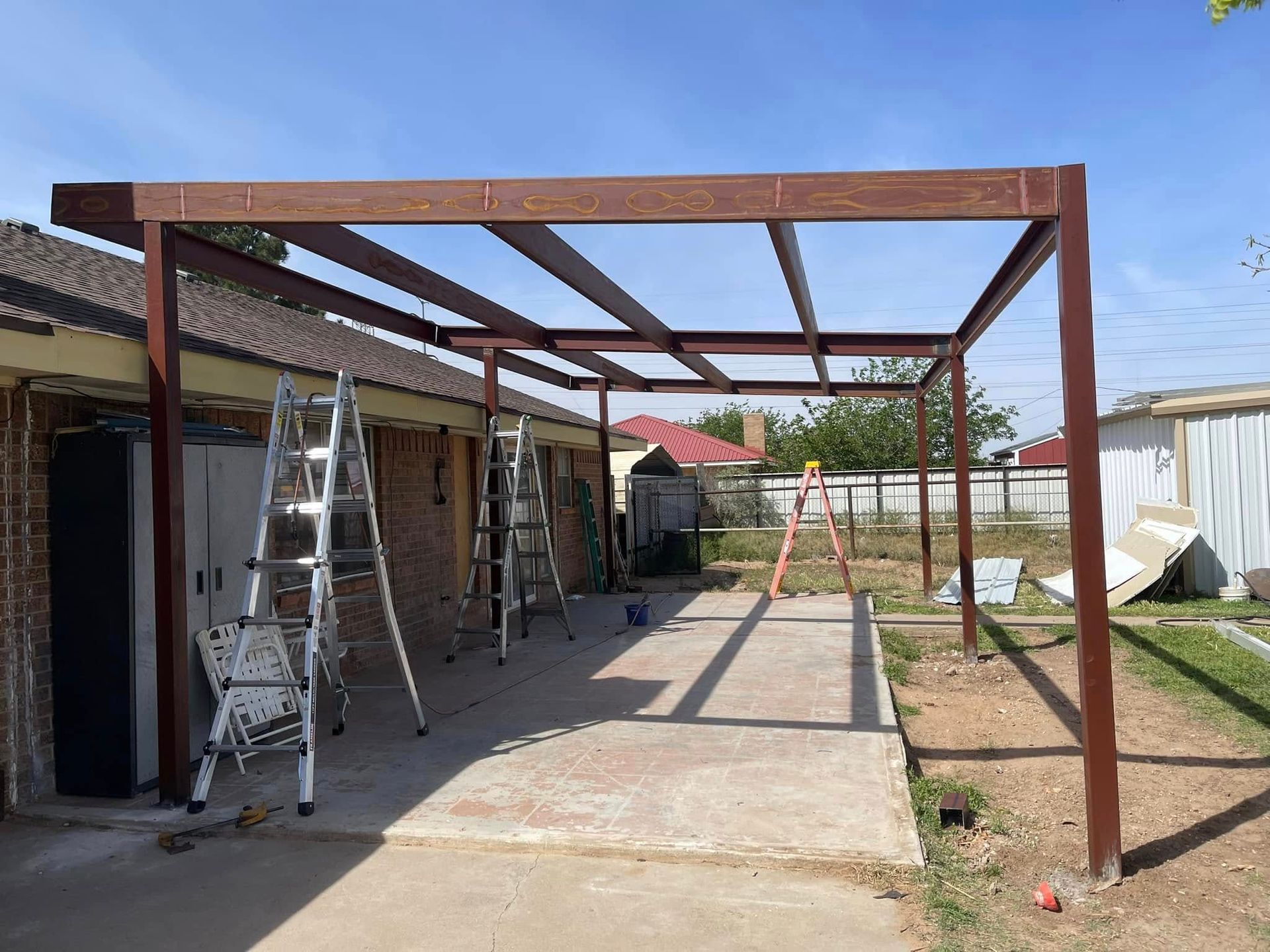 Partially built metal patio frame beside a house, with ladders and construction tools in a sunny yard