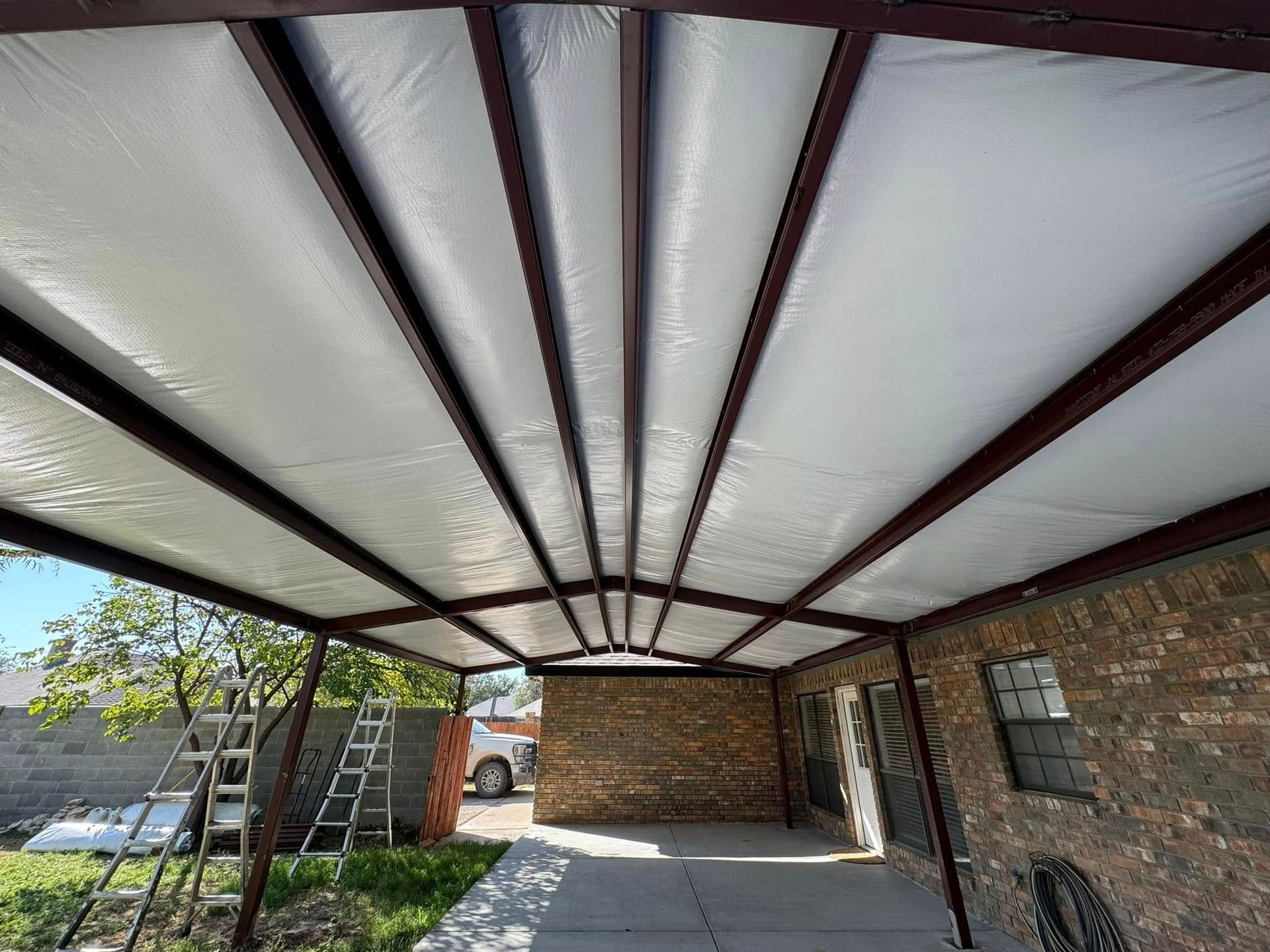 Covered driveway/carport with a white roof, dark beams, brick wall, and a ladder by a tree.