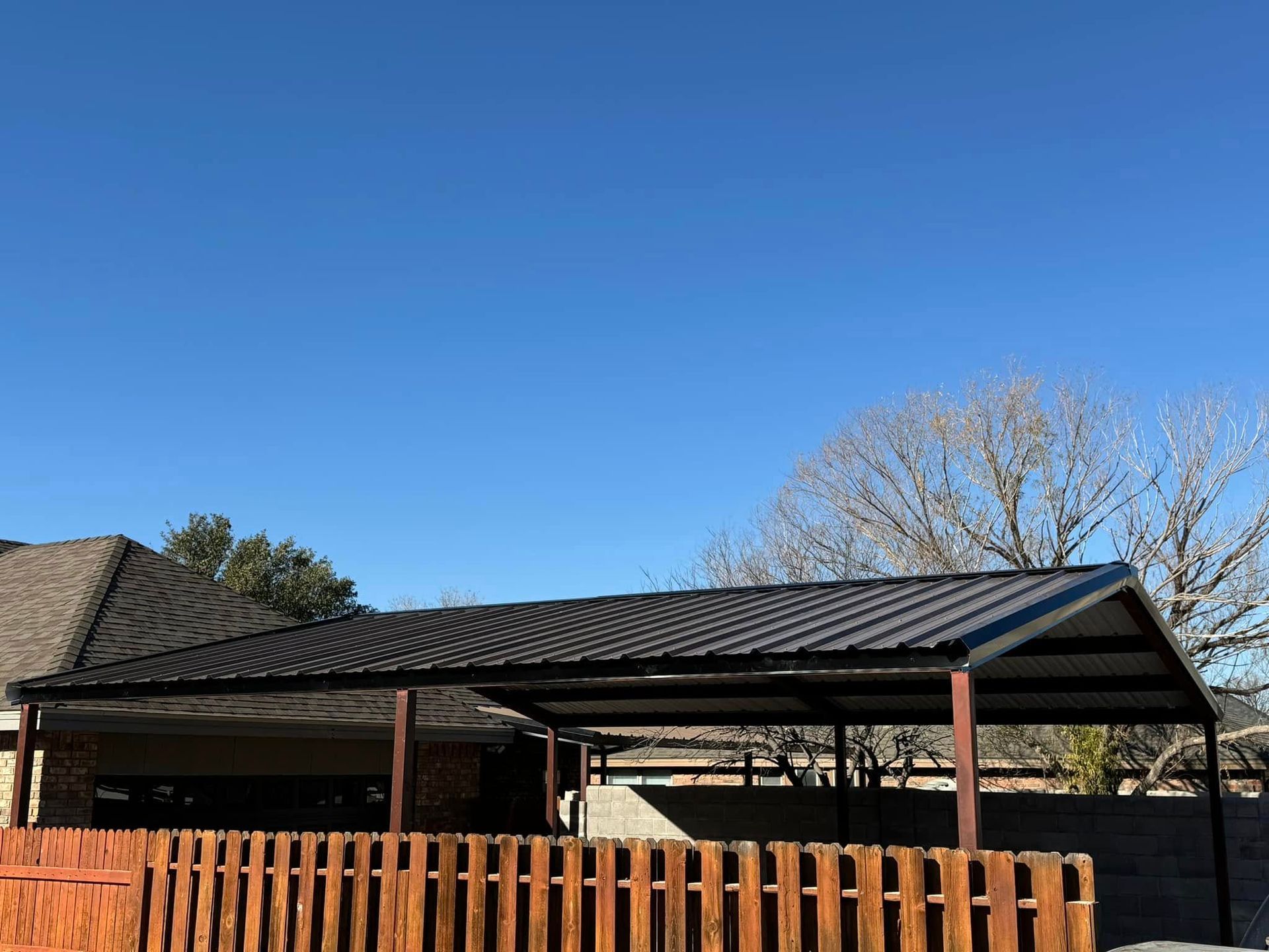 Backyard carport with black metal roof and wooden fence under a clear blue sky