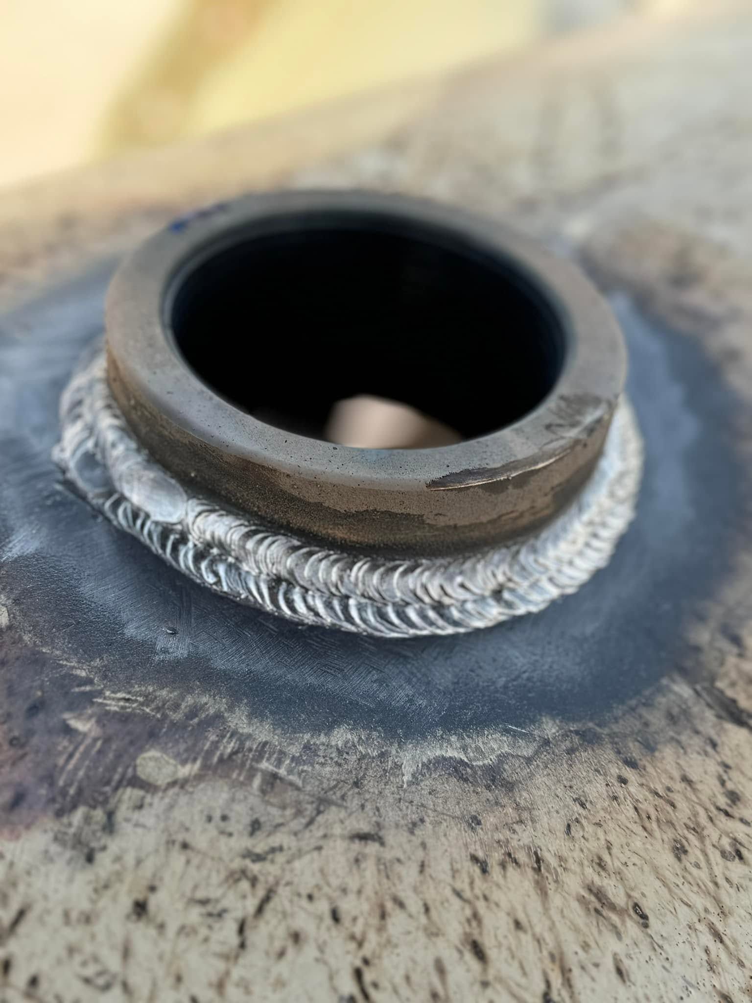 Close-up of a metal pipe opening welded onto a dark, textured surface with white residue around the seam