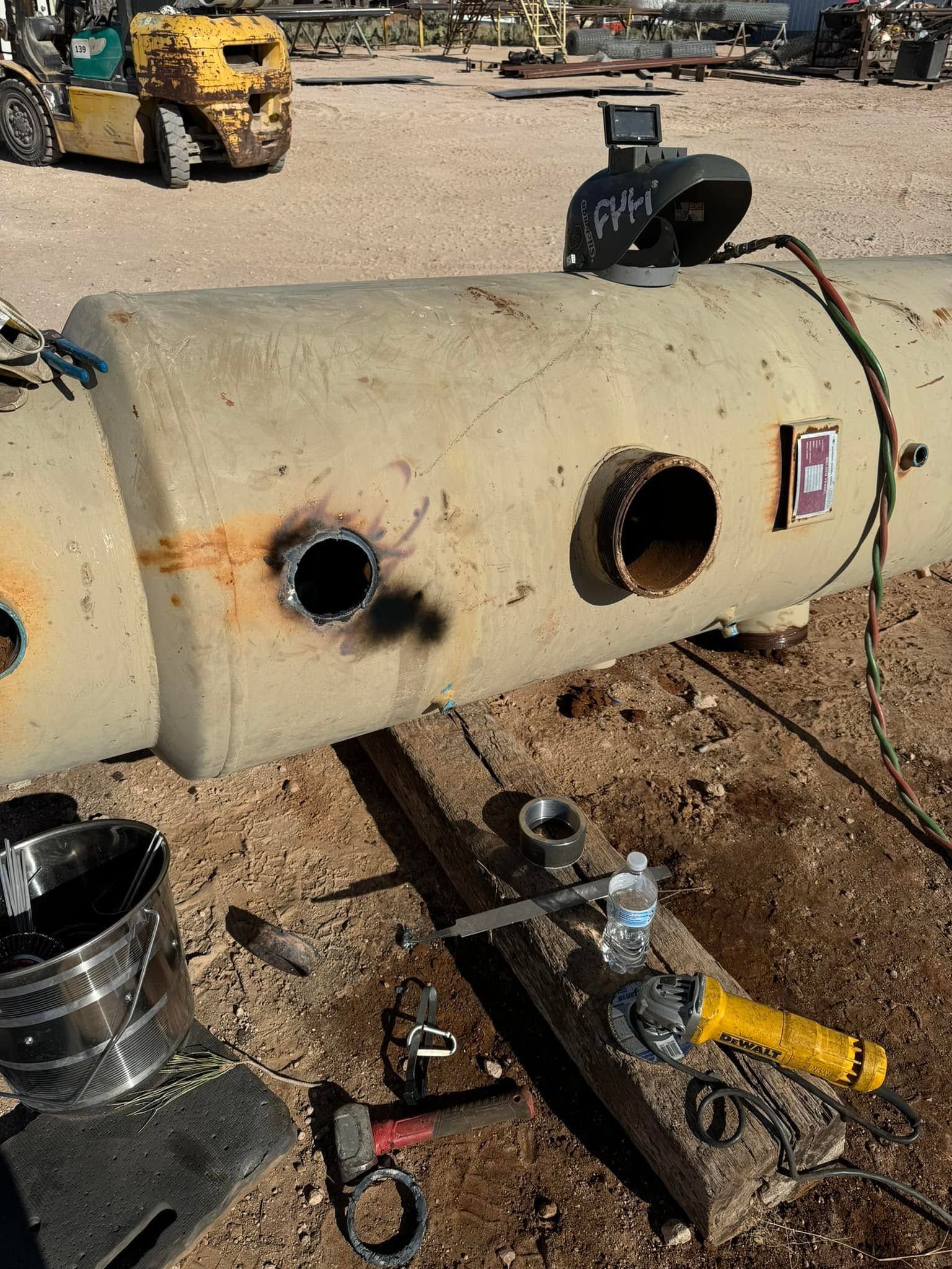 Large horizontal metal tank with two circular openings in a dusty worksite, tools nearby.