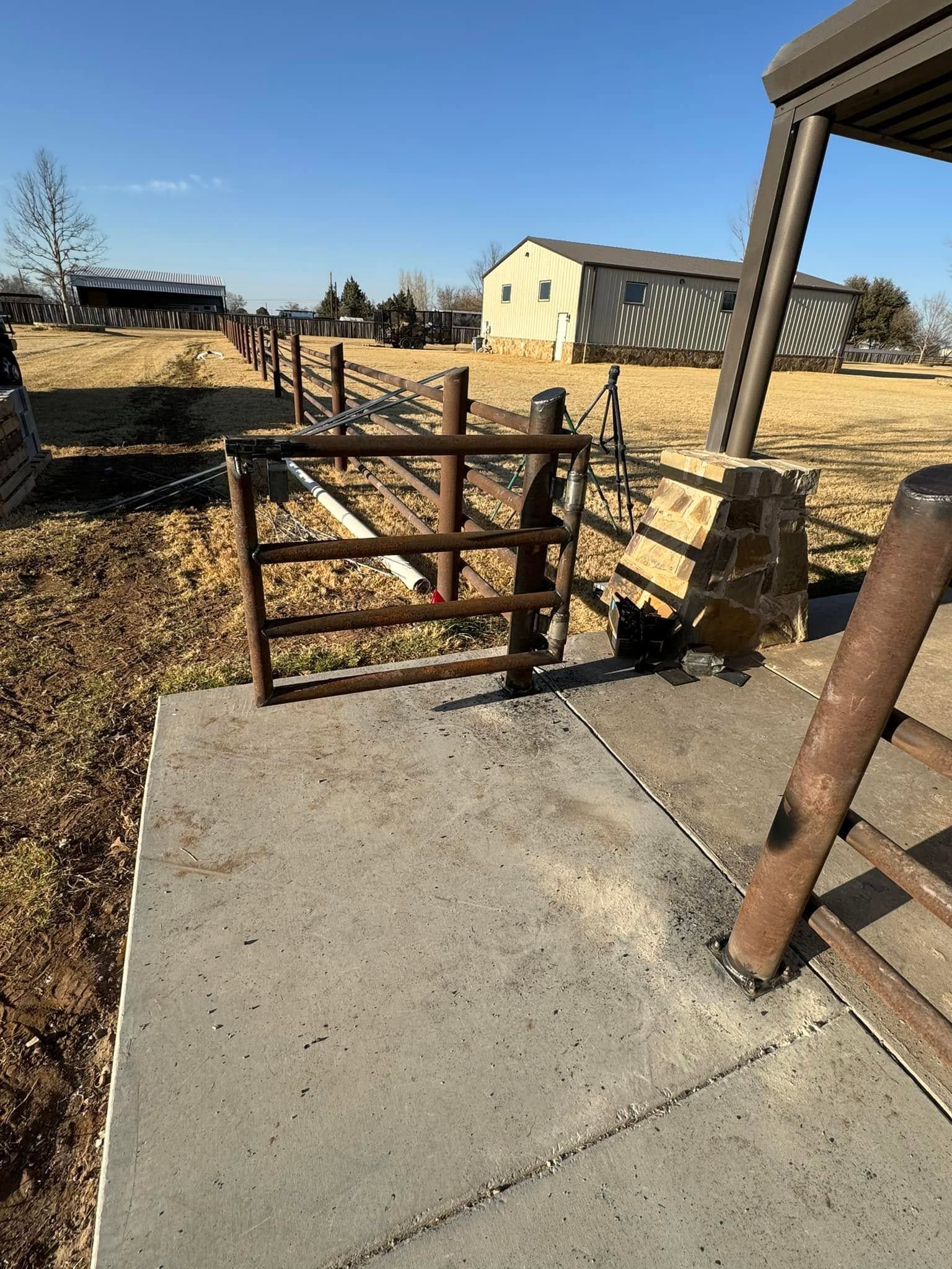 Rusty metal gate blocking a concrete path in a dry, open yard with a yellow house in the background
