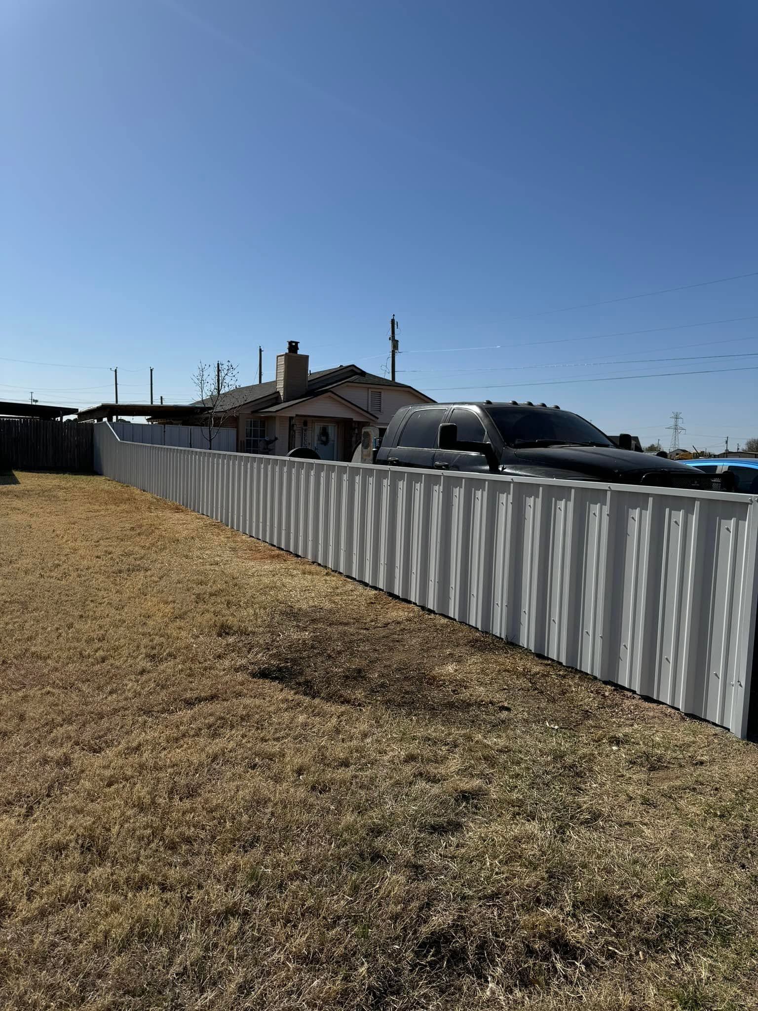 Long white fence beside a dry grassy field under a clear blue sky, with houses and cars in the distance