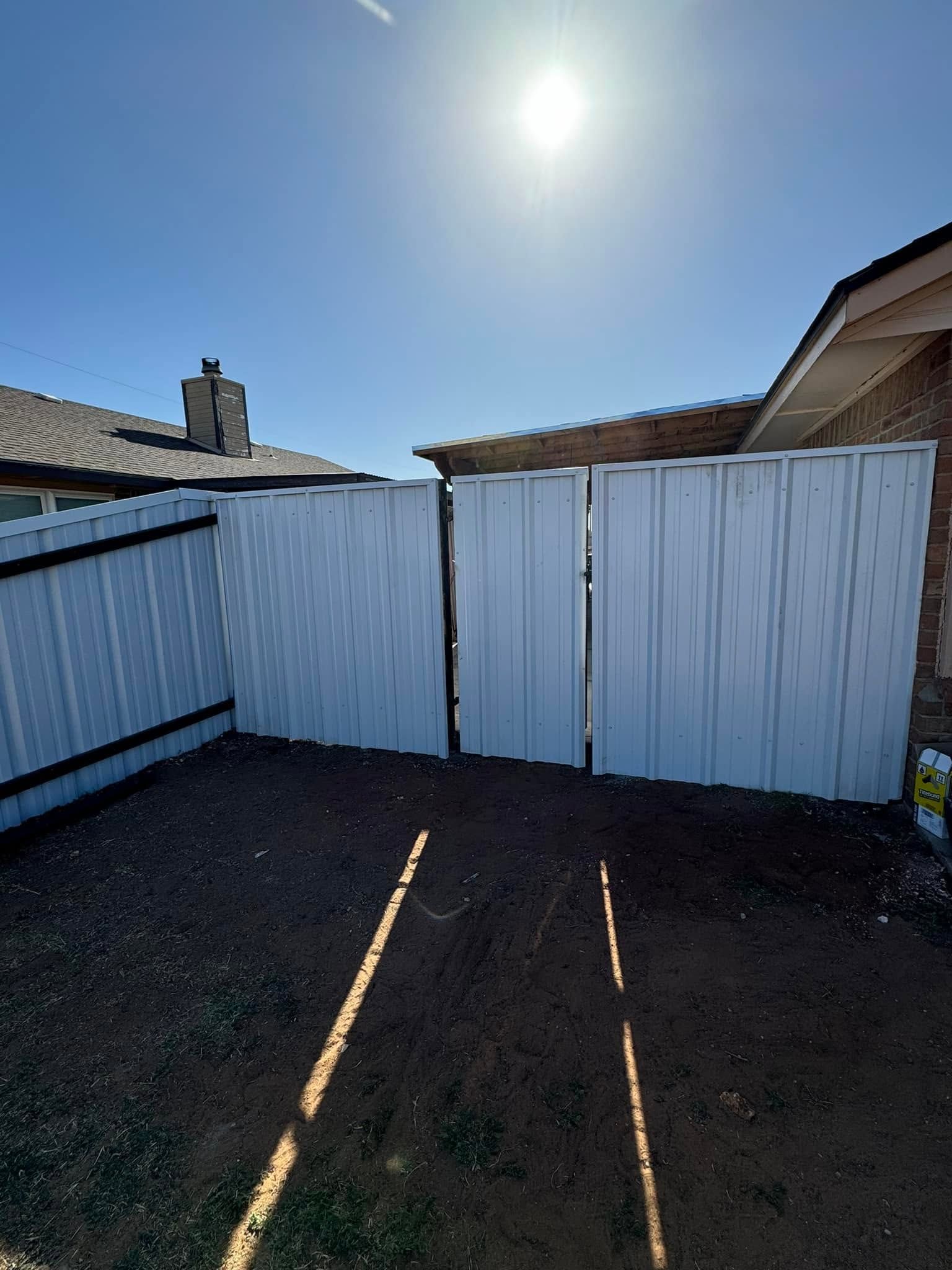 White wooden fence panels in a sunny backyard with a driveway and neighboring houses visible.