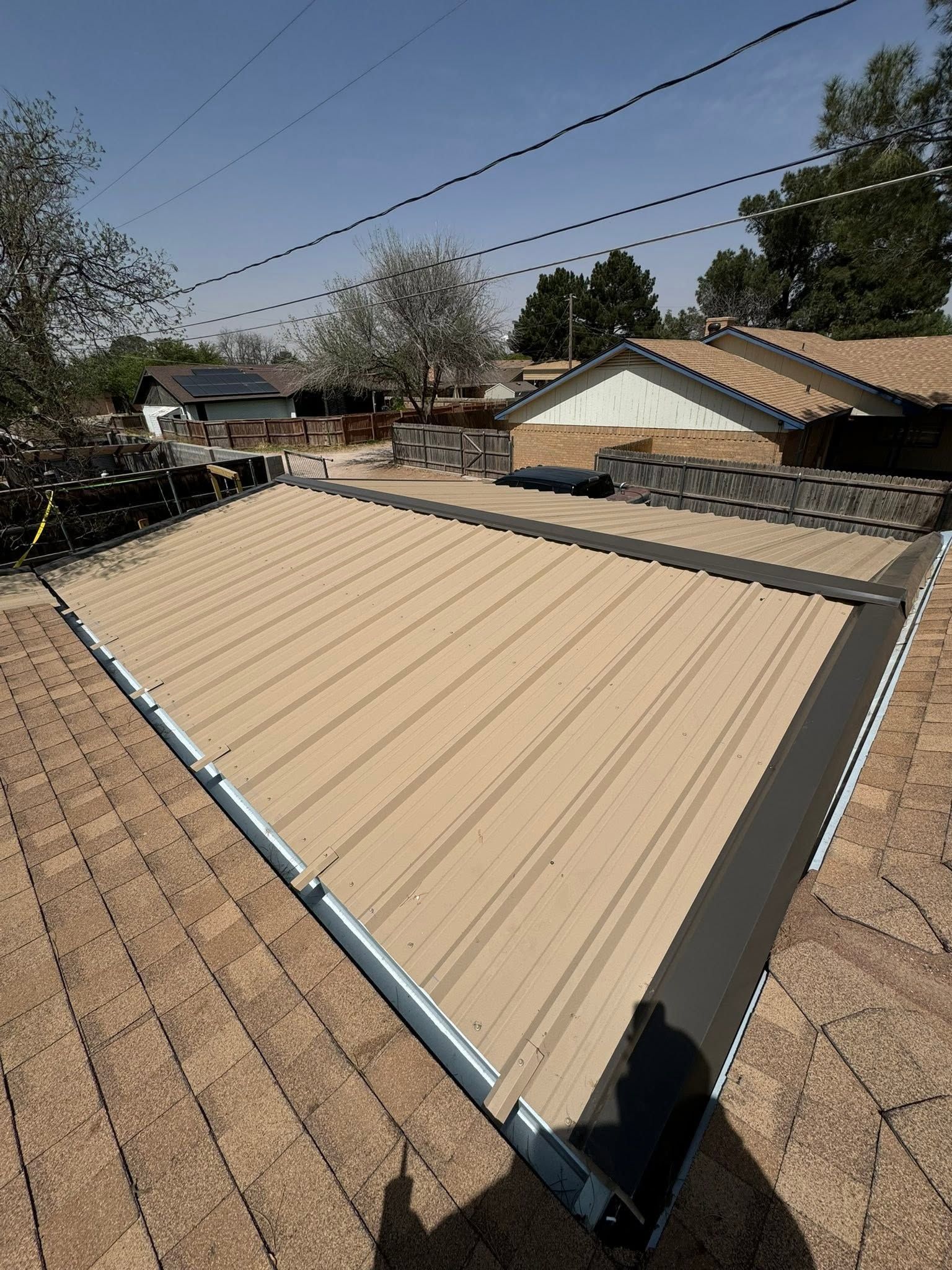 Tan metal roof with gray trim beside brown shingle roofs under a clear sky