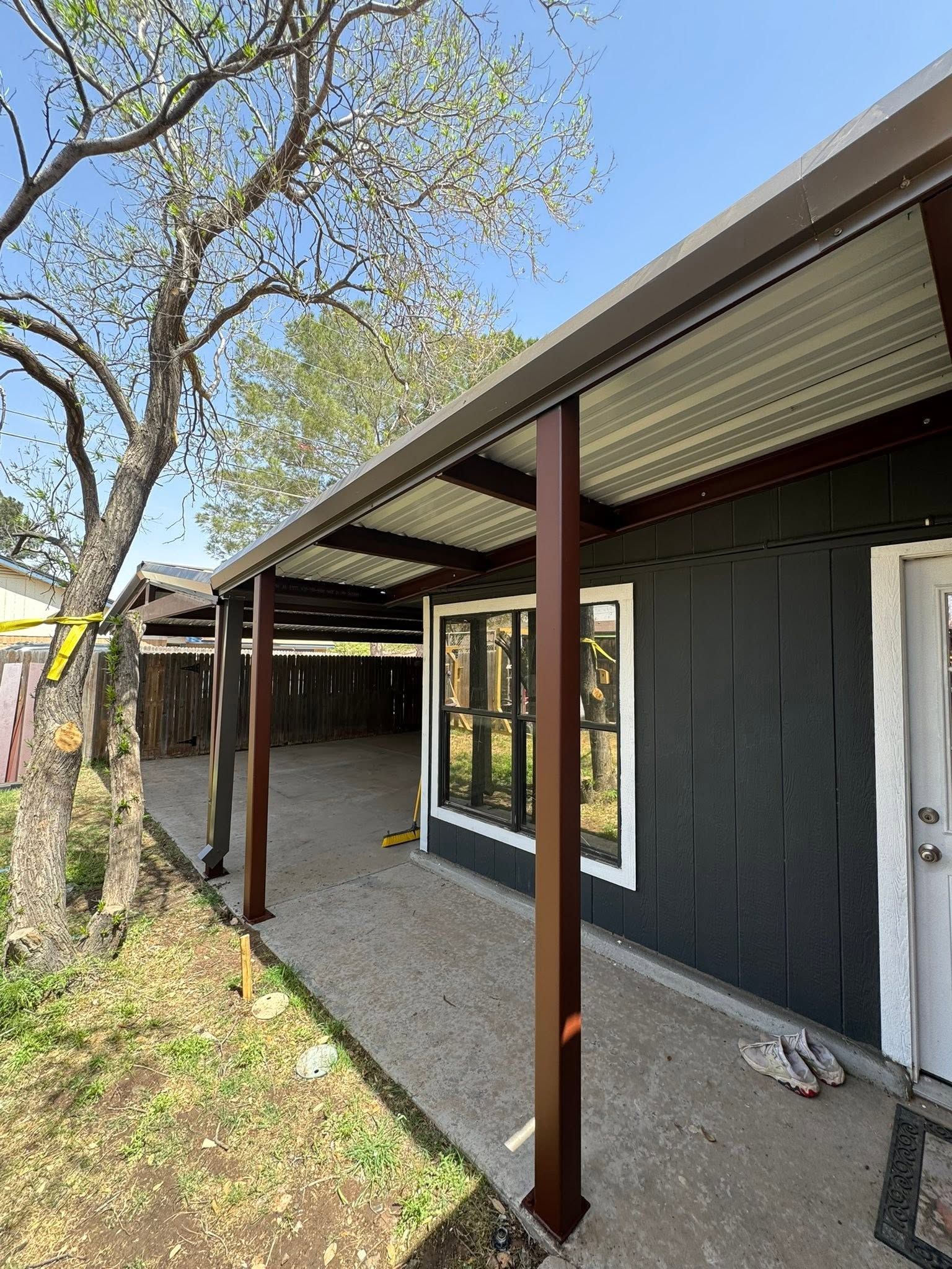 Covered walkway beside a house with brown posts, black siding, and a tree in the yard