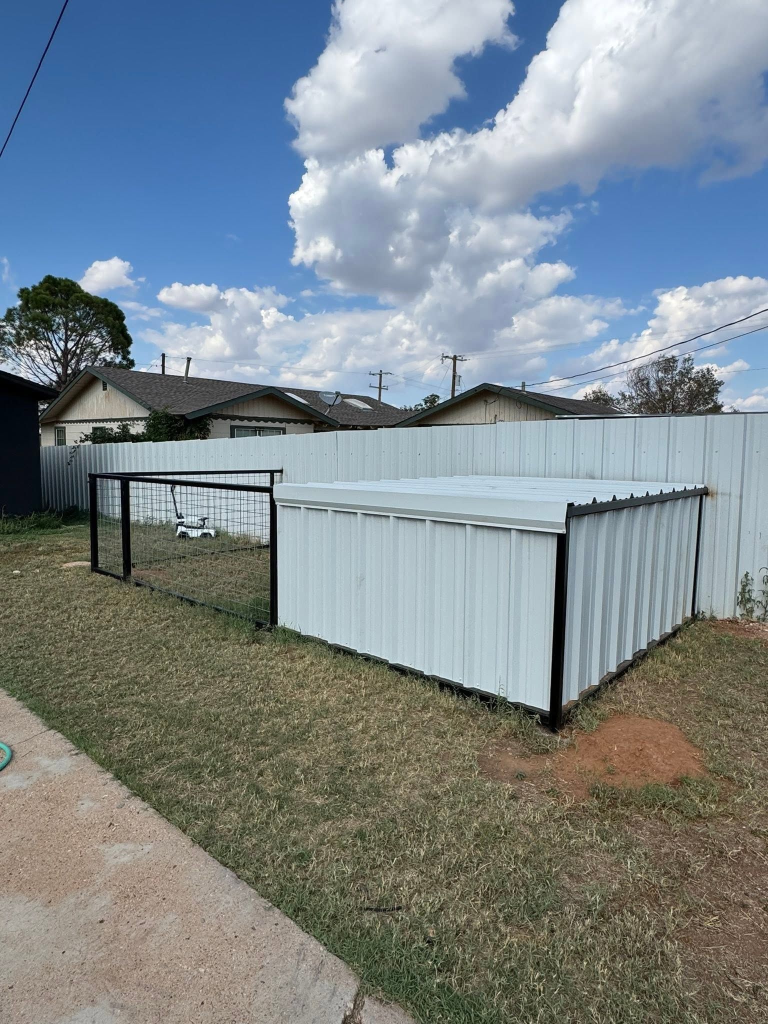 White corrugated metal sheds in a grassy yard under a partly cloudy sky, with houses in the background