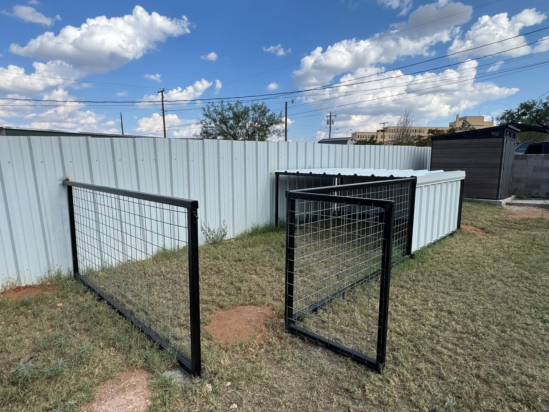 Open black metal gate panels in front of a white fence on a grassy yard under a cloudy sky