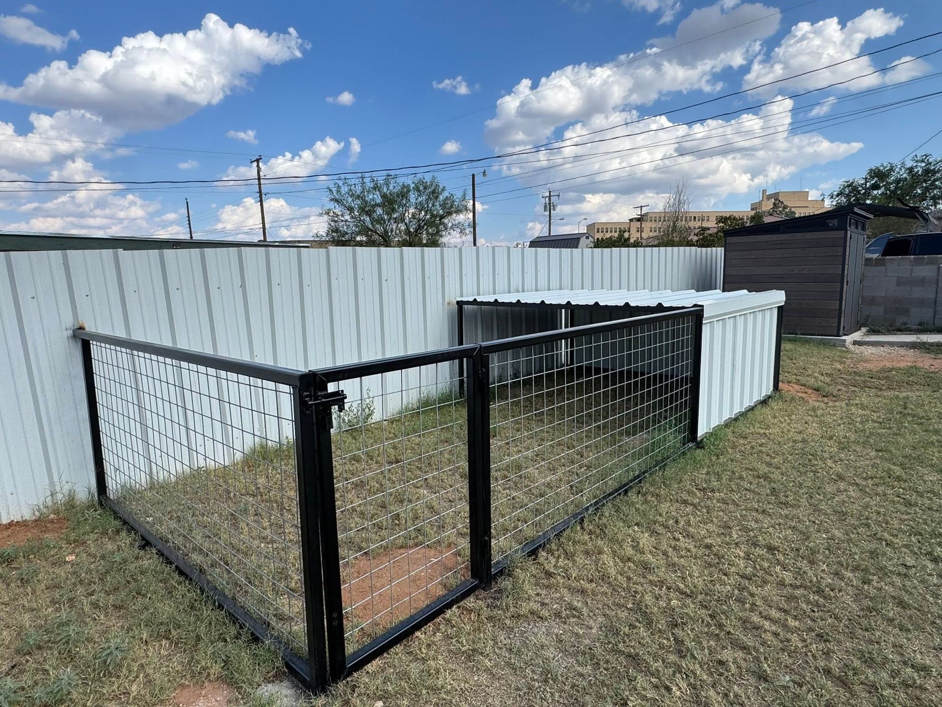 Black metal fence panels leaning beside a white corrugated fence in a grassy yard under a blue sky.