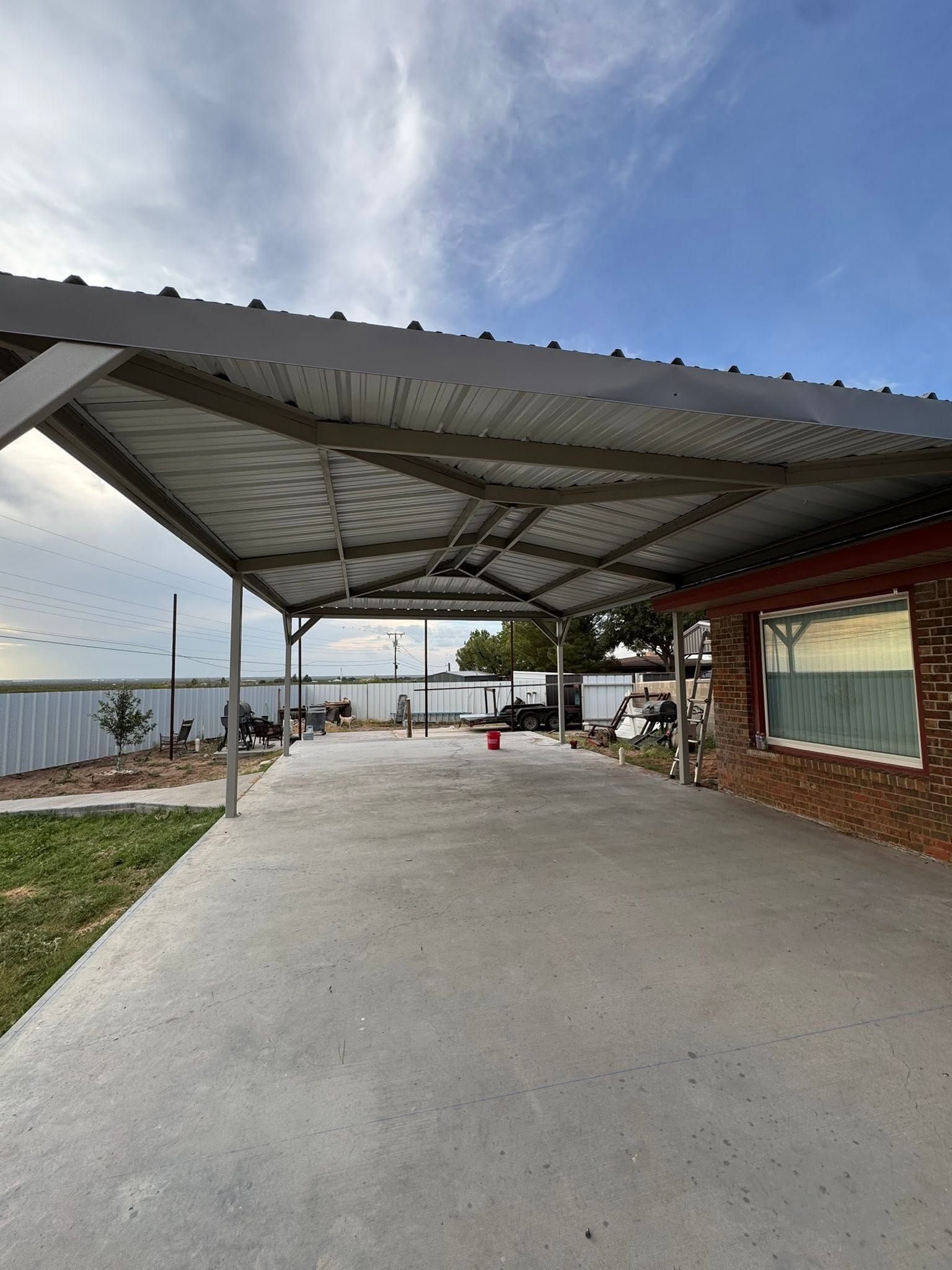 Covered concrete patio beside brick house, with metal roof, open view, and cloudy sky.