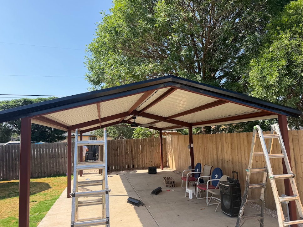 Open-sided metal carport with ladders and tools in a fenced backyard under construction