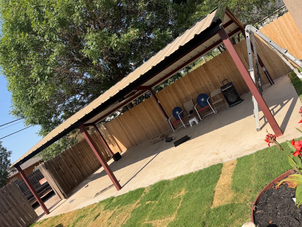 Outdoor covered patio with wooden posts, chairs, concrete floor, and a grassy yard beside a house.