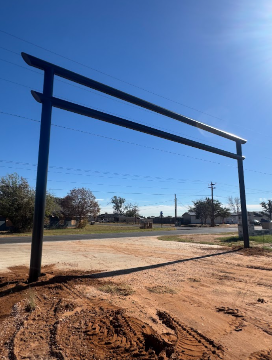 Tall black metal frame over a dirt lot under a clear blue sky, with a road and trees in the background