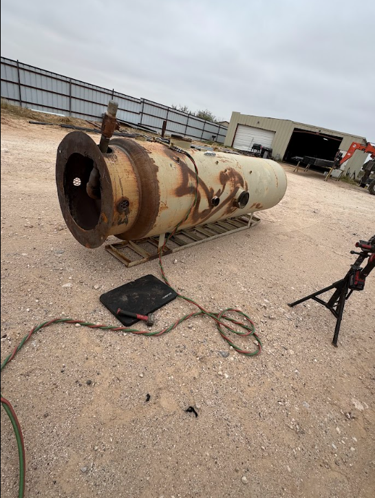 Rusty cylindrical tank on gravel near a fence, with a cable and tripod in an industrial yard