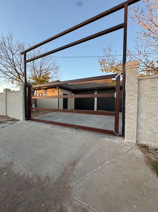 Tall brown metal gate between stone walls, closed across a residential driveway at dusk.