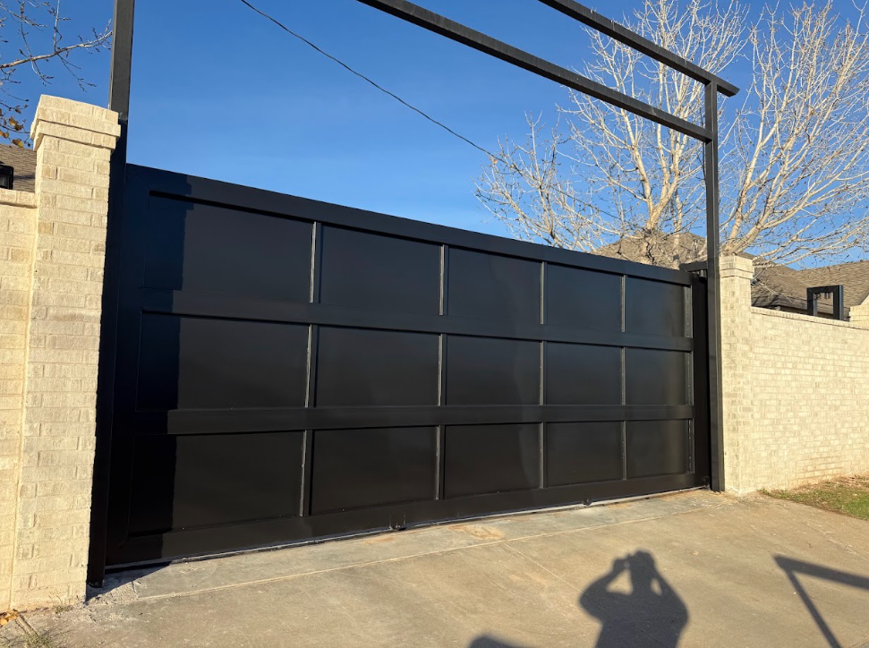 Black metal sliding driveway gate between beige stone walls under a clear blue sky