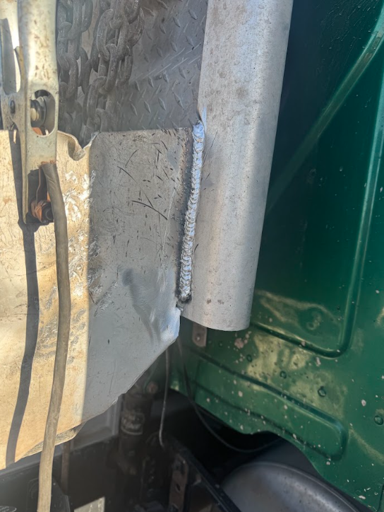 Close-up of a damaged gray and green vehicle panel with cracked paint and exposed metal, near a tire.