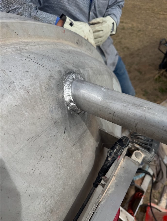 Person welding a metal pipe to a large gray tank, with sparks at the joint.
