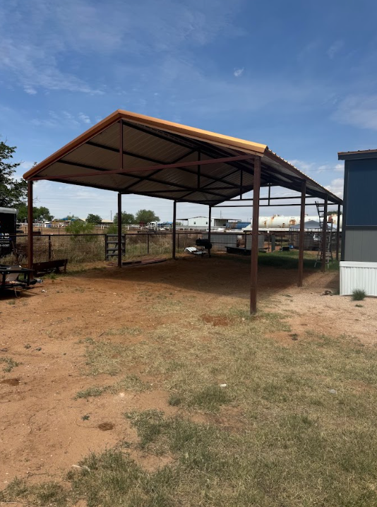 Open-sided metal pavilion with a corrugated roof on a dry dirt lot, beside a building and chain-link fence.