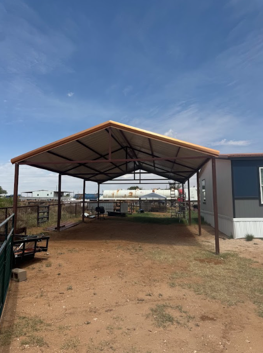 Open-sided metal shed on a dirt lot under a blue sky, with a building attached on the right.