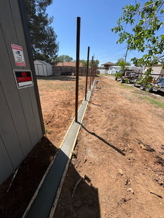 Narrow dirt path beside a fence and shed, with parked cars and trees in a sunny yard