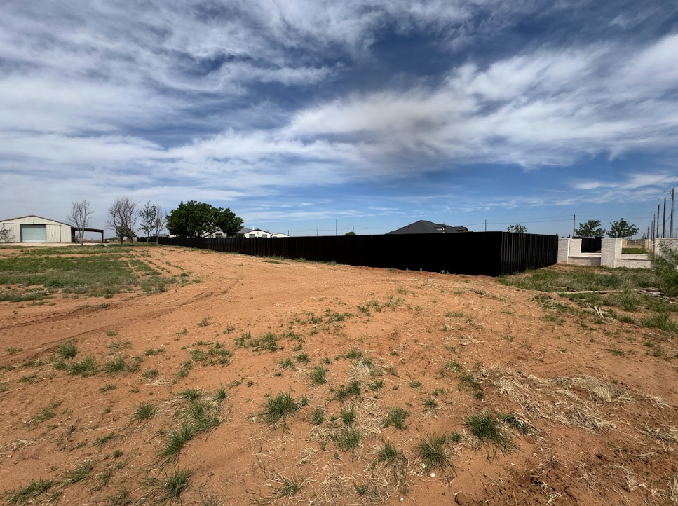 Dirt lot with sparse grass, black fence, and small buildings under a cloudy sky