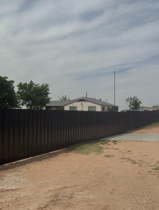 Rural dirt road beside a long black fence and a small house under a cloudy sky