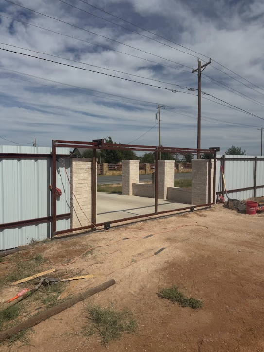 Partially open metal gate and fence at a dusty construction entrance under a cloudy sky