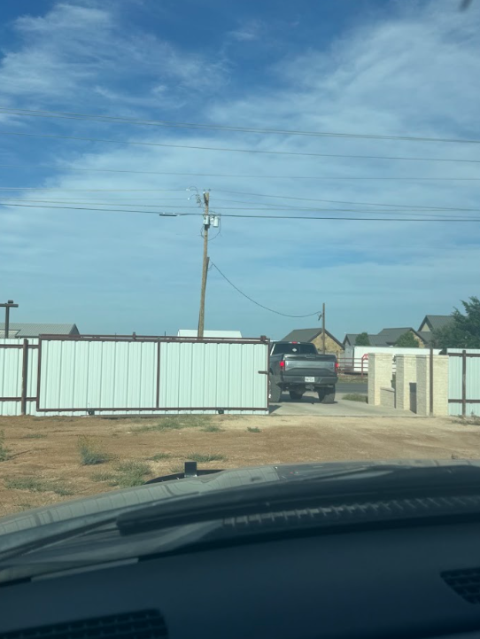View through a car windshield of a dirt driveway between white fences and a utility pole under a blue sky